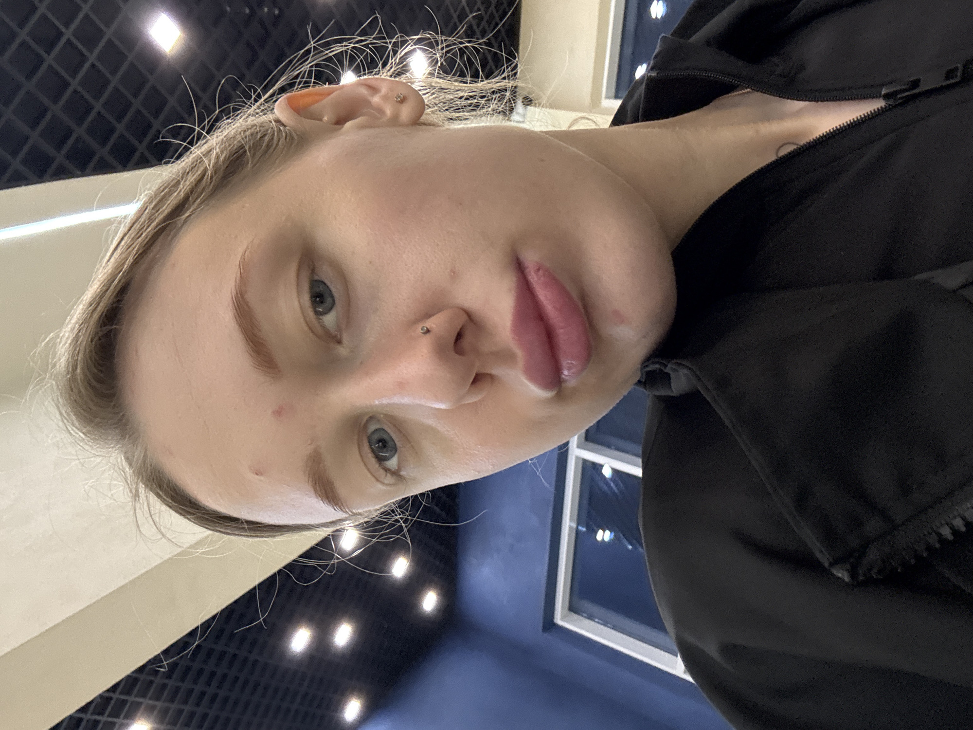 A close-up, slightly rotated portrait shows a woman with fair skin and light blonde hair, illuminated by artificial lighting. She has blue eyes and piercings in her nose and ear. She is wearing a black jacket, partially zipped, revealing a small circular tattoo on her neck. The background suggests an indoor setting, possibly a commercial space or an office, with a grid of lights on the ceiling and a window. There's no specific information to place the location in Rostov-on-Don.