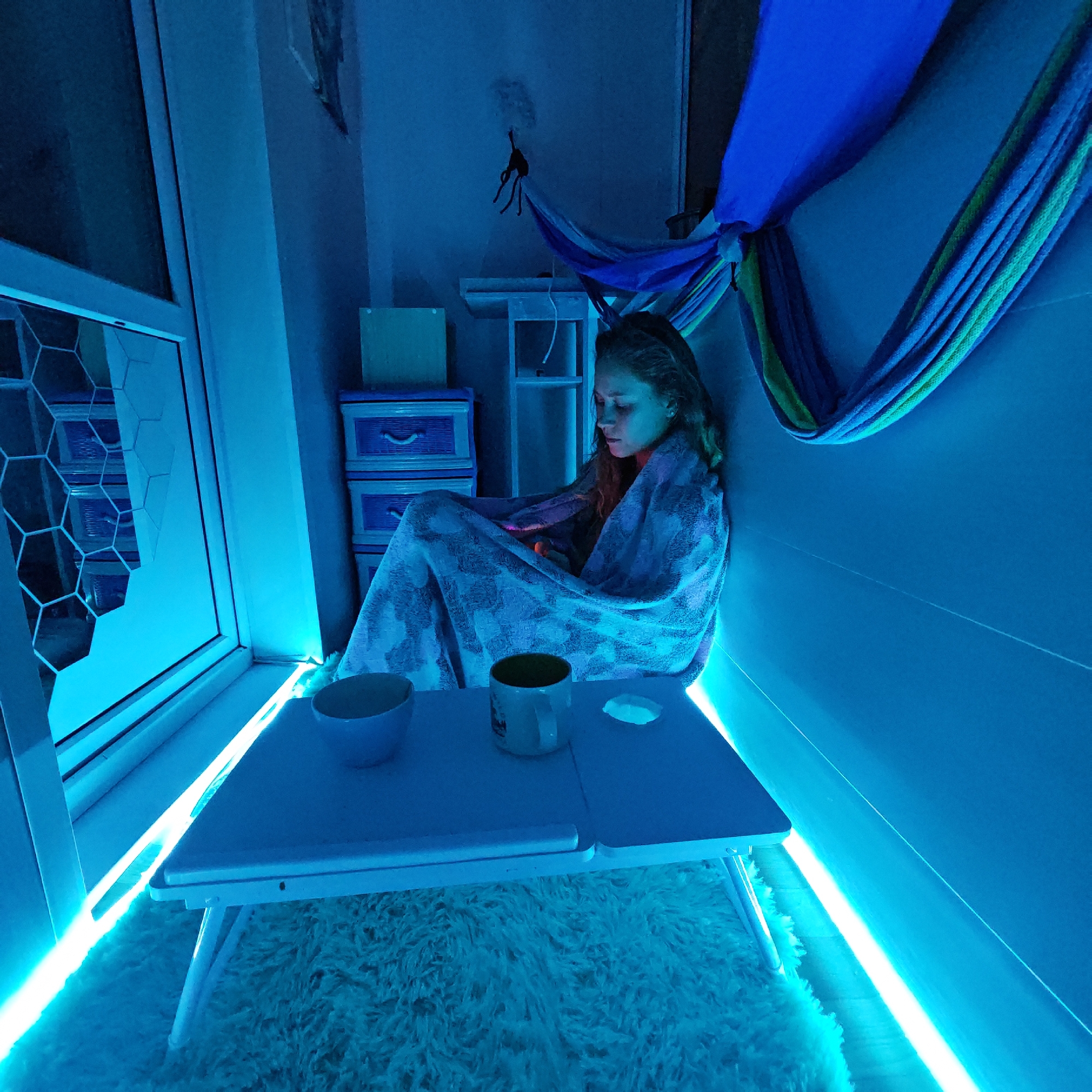 A young woman is seated in a small, confined space, possibly a balcony, in Rostov, Russia. She is wrapped in a blanket and appears to be looking down at something in her lap. In front of her is a small white table with a bowl, a mug, and a small white object on it. The room is illuminated with a strong blue light, likely from LED strips, which highlights the white shag rug underneath the table. A window with a geometric pattern is visible to the left. Behind her, a colorful hammock-like fabric is suspended. Stacking plastic drawers are stacked behind the woman. A small toy is fixed to the wall.