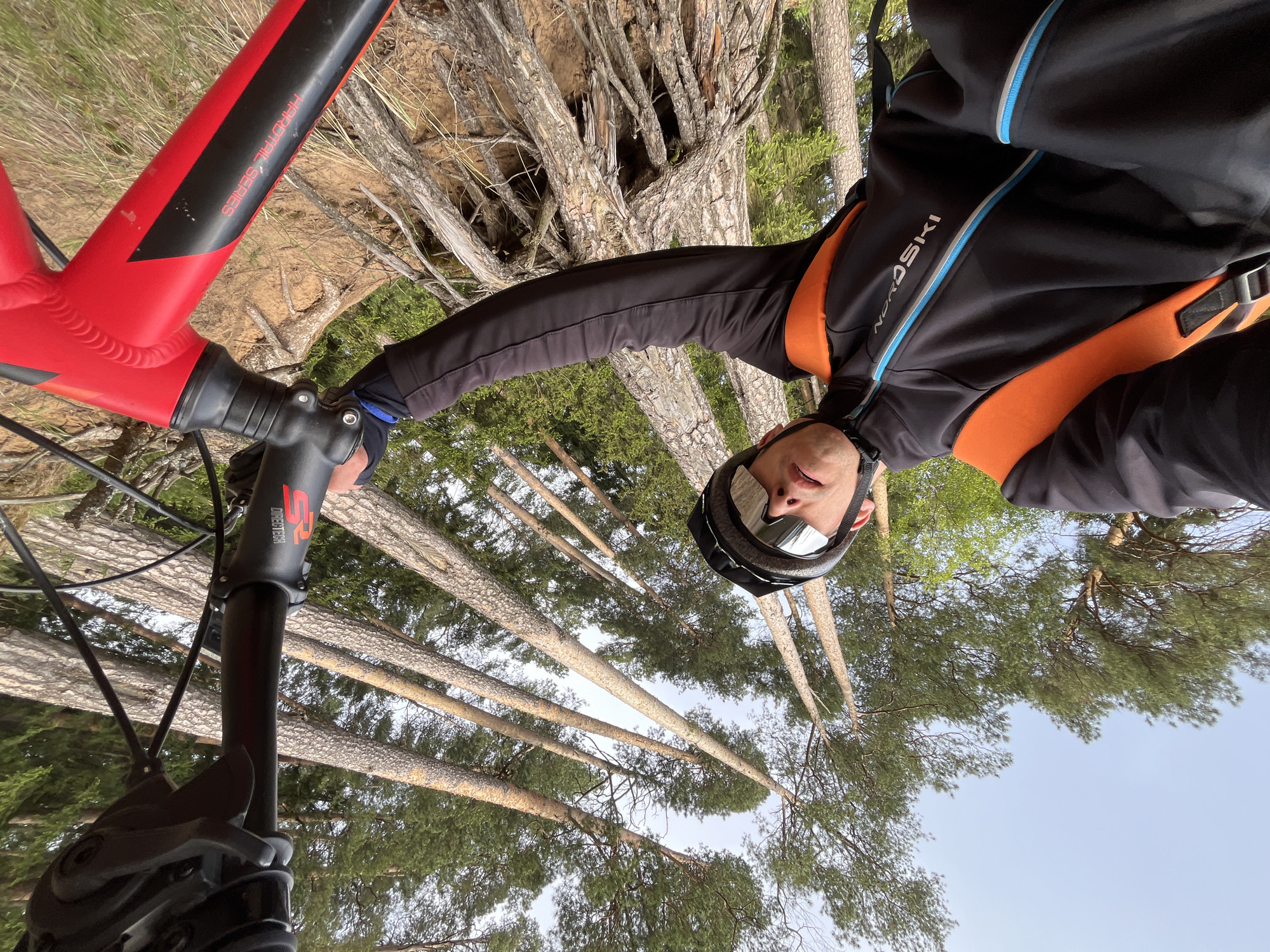 A low-angle, medium shot depicts a person on a red and black mountain bike in a forest. The person, wearing a black helmet with reflective goggles and a black and orange jacket with the word "NORD SKI" printed on the side, is seen from the chest up. The person's face is visible, looking directly at the camera. The bike is centered on the left side of the image. The handlebars are black, with the inscription "SR SUNTOUR" on the stem. Behind the person and bike, a dense forest stretches towards the sky, with tall, thin trees and green foliage. The location is likely in the vicinity of Chaikovsky, Russia, judging from the person's attire and the environmental context.