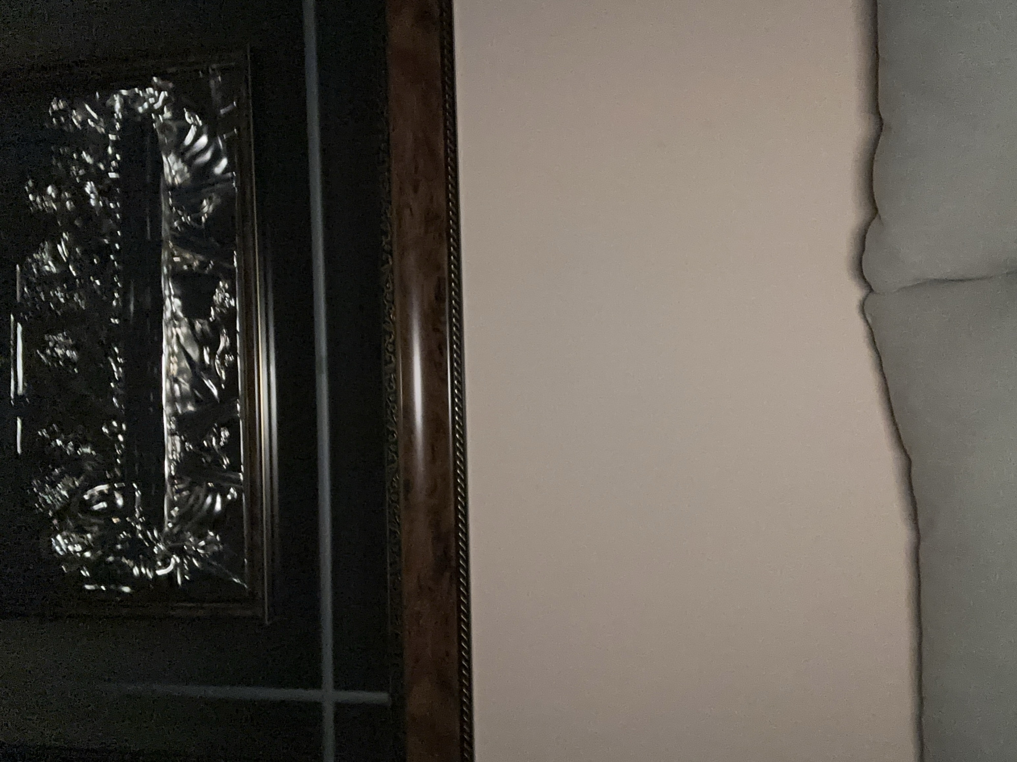 The image shows a close-up of a dark, ornate wooden door or cabinet with a silver-colored decorative element. The doorframe is also made of dark wood with a decorative trim. To the right of the door, there is a light pink wall, with a textured edge suggesting a corner or a different material. The lighting is dim and appears to be coming from an unknown source. There is no information to identify the location as Yerevan, Armenia, or to indicate any people or actions taking place.