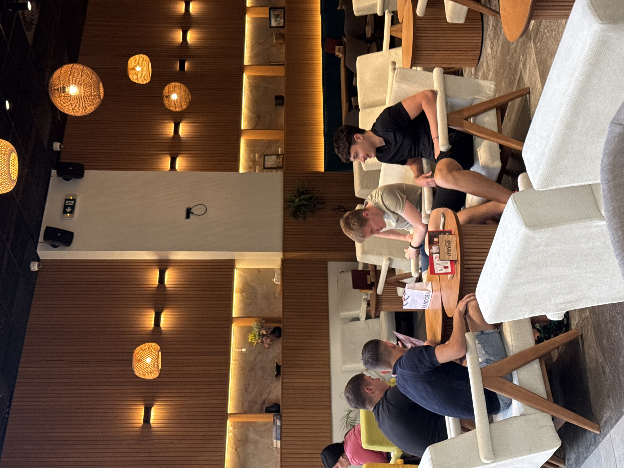 Several people are seated at a table in a cafe in Belgrade, Serbia. A mix of indoor lighting fixtures are present. The cafe has wooden paneling and shelves with decorative items. Some of the individuals are engaged in conversation, while others are focused on their phones. The cafe appears to be in a contemporary style, with white upholstered chairs and wooden tables.