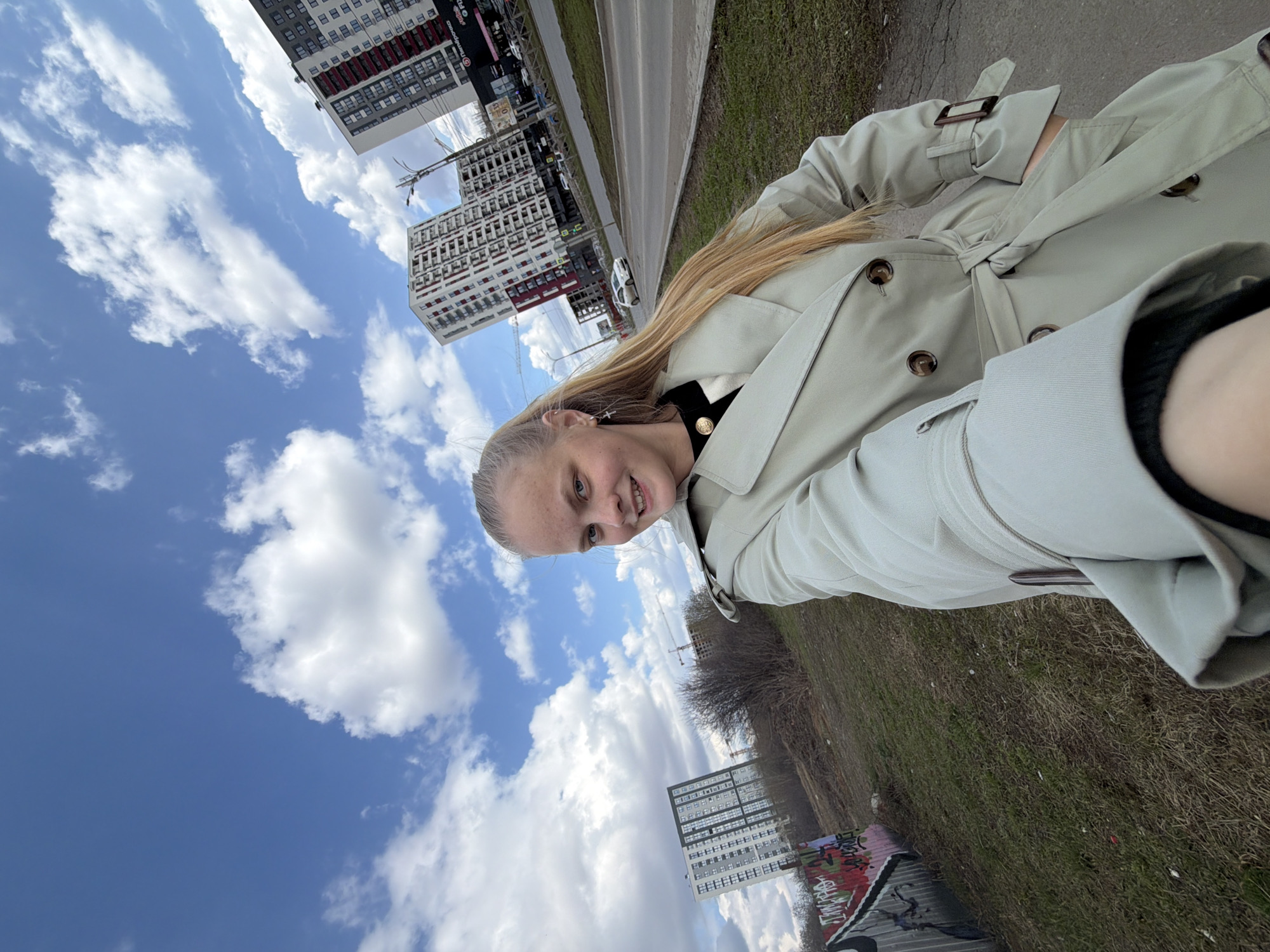 A young woman is visible in a selfie-style photograph taken outdoors, seemingly near an urban environment. She has fair skin, blond hair, and is wearing a beige trench coat. Her face is visible, and she appears to be smiling. The background shows a blue sky with white clouds, several high-rise apartment buildings, and some greenery along a roadside. There is a road with cars, and a sidewalk near the woman. Graffiti is visible on a structure near the bottom of the photograph. This appears to be Izhevsk, Russia.