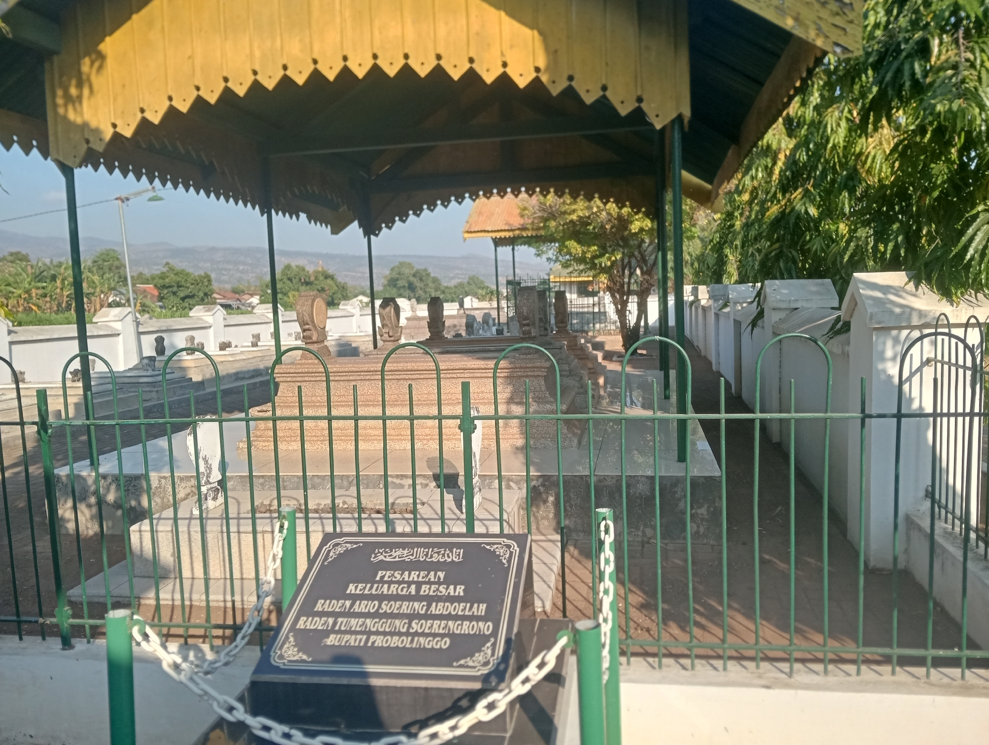 The image shows the grave site of the Raden family in the city of Bloro, Indonesia. The grave is enclosed by a green metal fence and a white stone wall. A yellow-roofed pavilion covers a portion of the grave site. A dark plaque with white text, in Arabic and Indonesian, is visible in the foreground. The plaque reads "PESAREAN KELUARGA BESAR RADEN ARIO SOERING ABDOELAH RADEN TUMENGGUNG SOERENGRONO BUPATI PROBOLINGGO." The background shows trees, houses, and hills. The lighting suggests it is either morning or late afternoon. There are no people visible in the image.