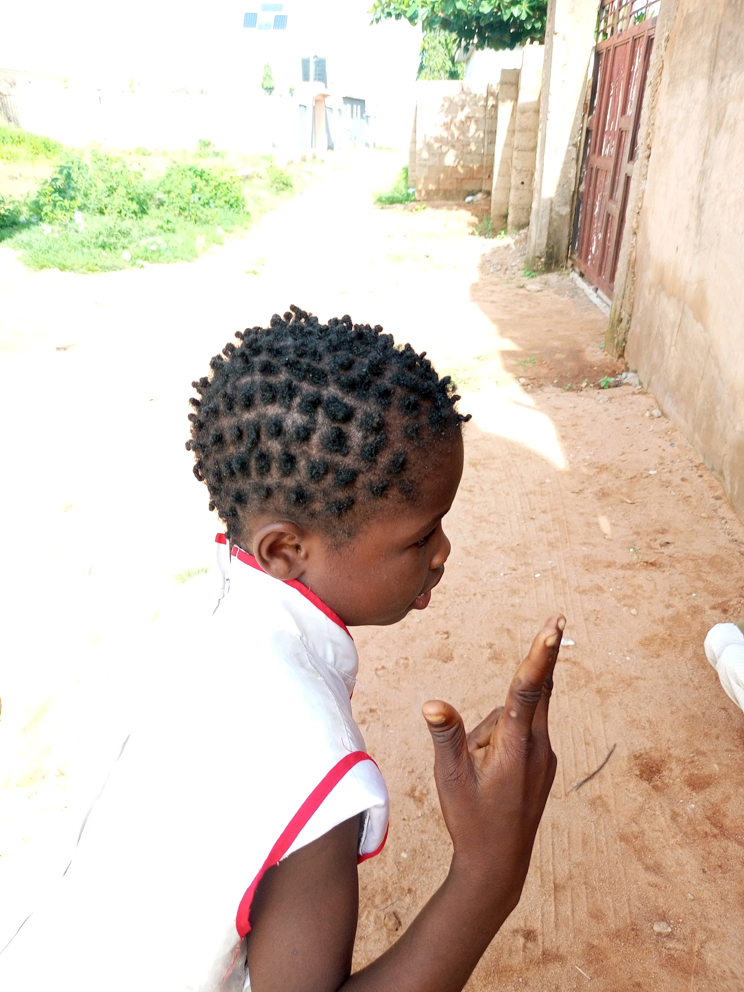 A young Black child, likely female, is positioned in the foreground of the image, facing right and slightly away from the camera. The child has short, dark, braided hair, styled in small, square sections. They are wearing a white garment with red trim around the neckline and armholes. The child's right hand is raised, with the index finger extended and the thumb bent.
The background depicts a dusty, dirt road in an outdoor urban setting. To the right, a beige-colored wall runs along the side of the road, featuring a reddish-brown metal gate with an intricate lattice design. Further down the road, stone pillars and a wall constructed of rough stones are visible. Vegetation, including green bushes and trees, borders the left side of the road, with a building or two faintly visible in the distance. The lighting suggests it is daytime, with shadows cast on the ground. The environment appears to be consistent with an area in Gombe, Nigeria, given the architecture, vegetation, and road conditions.