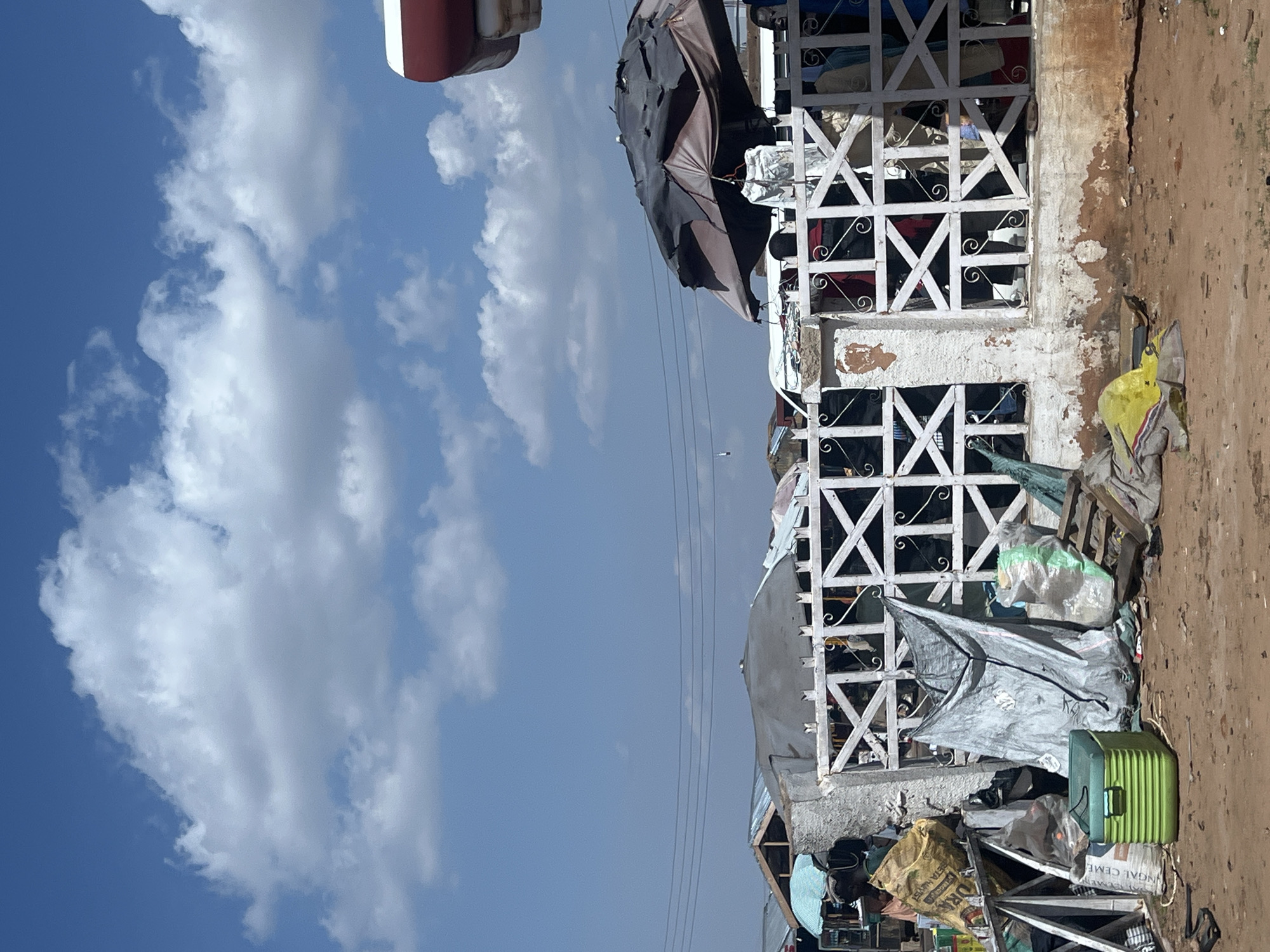 This image captures an outdoor market scene in Gombe, Nigeria, under a clear blue sky with scattered white cumulus clouds. The ground is a reddish-brown dirt path, uneven and littered with various items. In the immediate foreground, a bright green ridged cooler, multiple plastic bags (including yellow, green, and grey varieties), wooden planks, and a large yellow sack labeled "NGA CEME" are visible. A low, weathered white wall topped with a decorative white wooden trellis-style fence runs horizontally across the mid-ground, featuring interconnected 'X' patterns and scrollwork. Behind this fence, several indistinct figures are partially obscured, suggesting activity within the market stalls. Above the fence, two large market umbrellas are prominent: a torn, dark-colored umbrella on the right and a light blue and white patterned umbrella on the far left. In the background, numerous makeshift structures with assorted roofs (tarps and corrugated metal) indicate a bustling informal trading area. Horizontal power lines span the mid-sky. A partial red and white structure is visible at the top right edge of the frame.