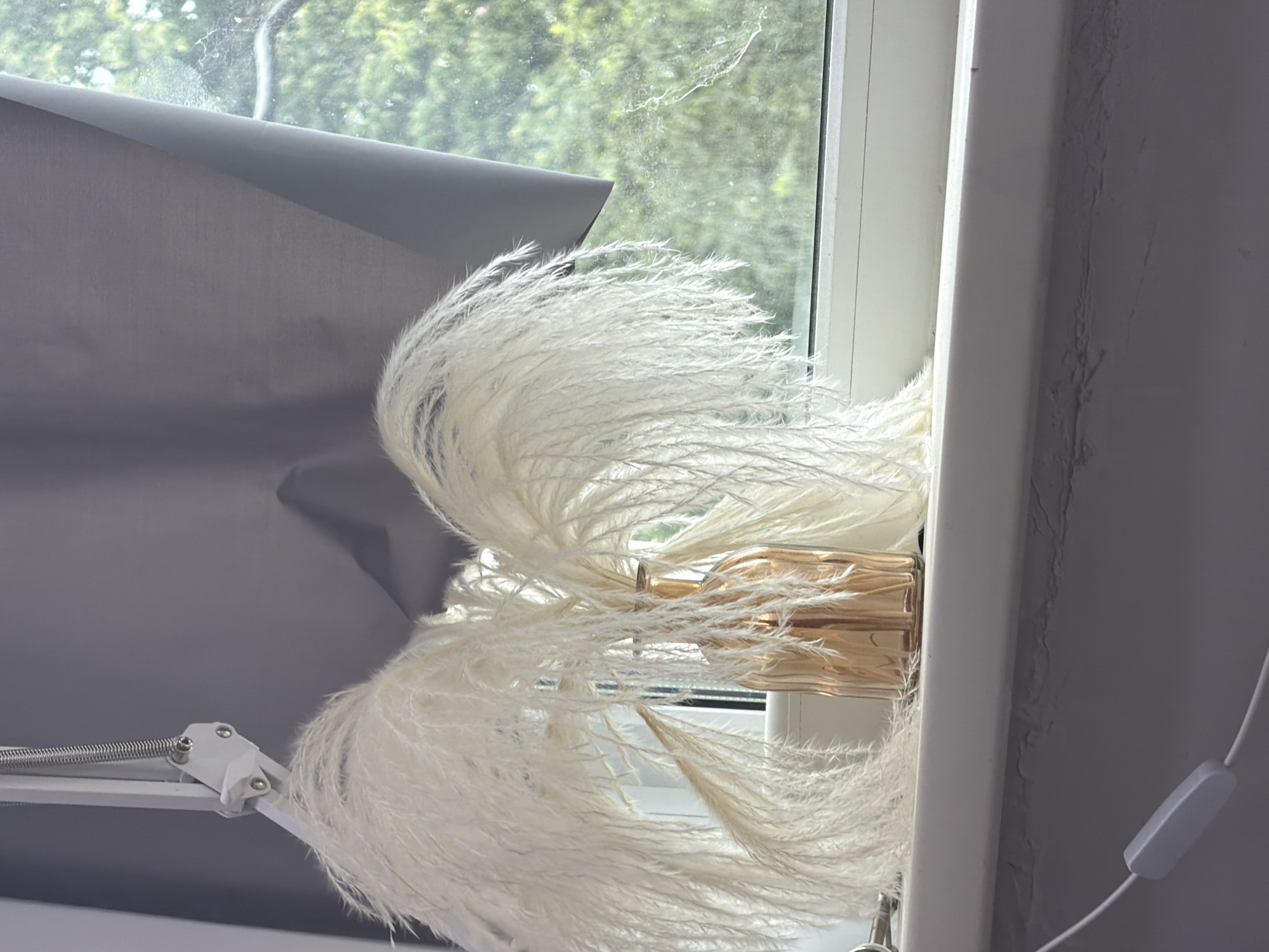 A close-up shot shows a copper-colored glass vase filled with tall, fluffy white reeds. The vase sits on a white windowsill, framed by a grey curtain on the left and a window with green foliage visible outside on the right. A white swing-arm desk lamp is partially visible on the left side of the frame, and a white electrical cord with an in-line switch runs along the bottom of the windowsill. The scene is lit by natural light coming through the window.