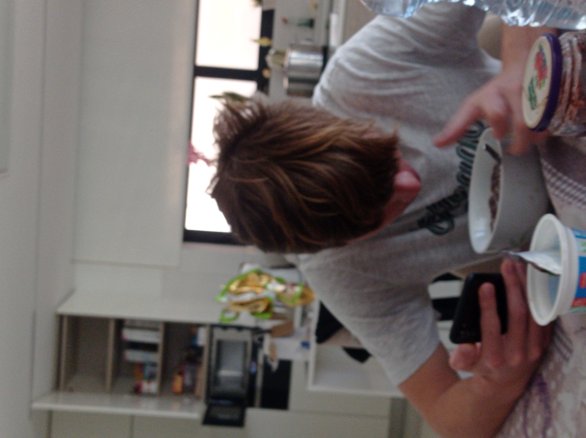 A person is seen indoors, possibly in a kitchen setting, holding a phone in one hand and two yogurt containers. A jar is also in the scene. The person is wearing a gray shirt. There's a window visible in the background and kitchen cabinets and appliances. No specific information suggests a location in Felgueiras, Portugal. No action or interaction is clearly taking place.