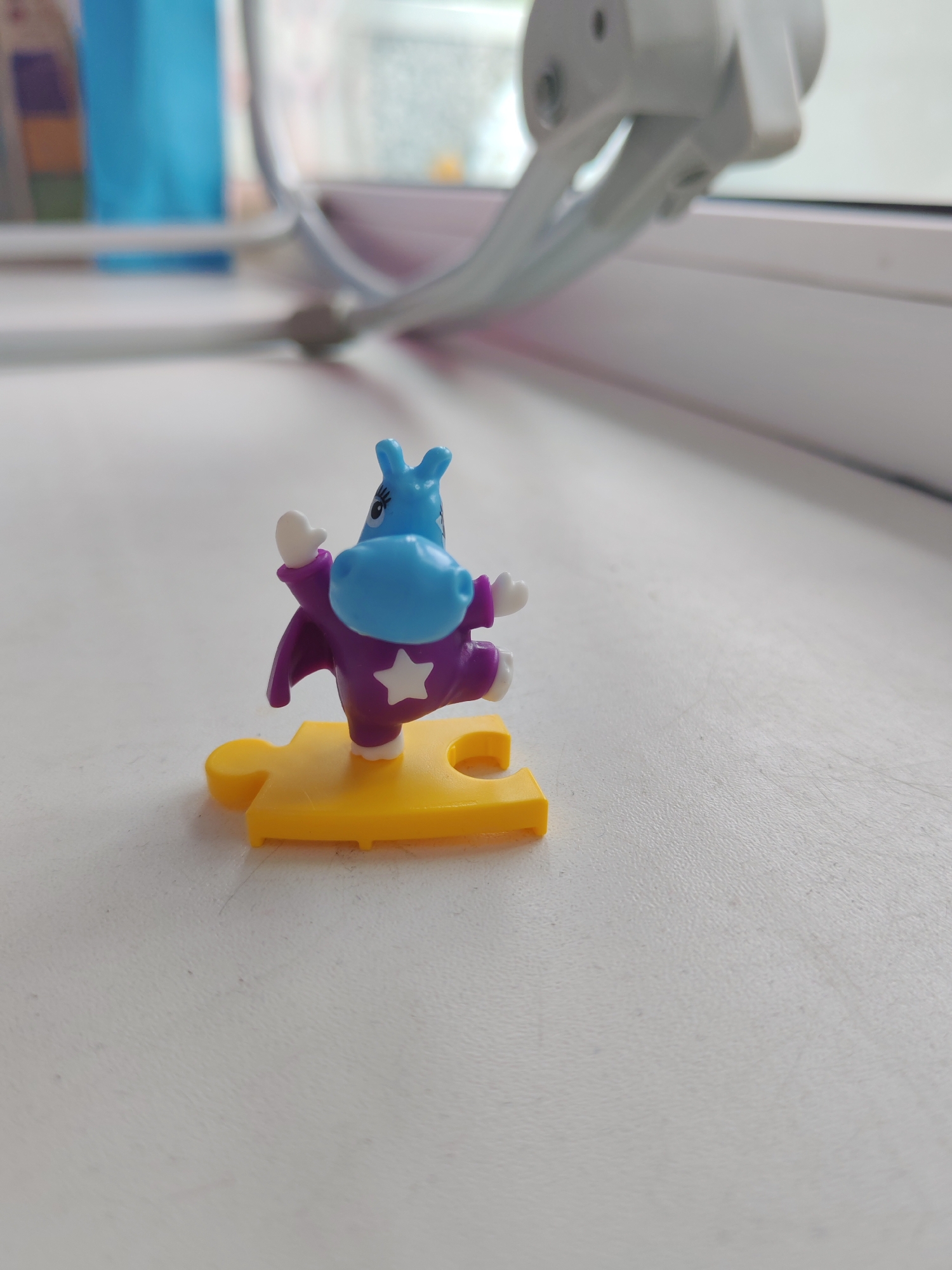 A small, blue plastic figurine of a hippopotamus wearing a purple cape with a white star is standing on a yellow puzzle piece. The figurine is positioned on a white surface, possibly a windowsill or desk. In the background, a blurred white and gray object, which appears to be a part of a window frame or blind mechanism, is visible. The overall scene is brightly lit, suggesting natural light from a window. The location in the city of Belogorsk, Russia cannot be determined from the provided image.