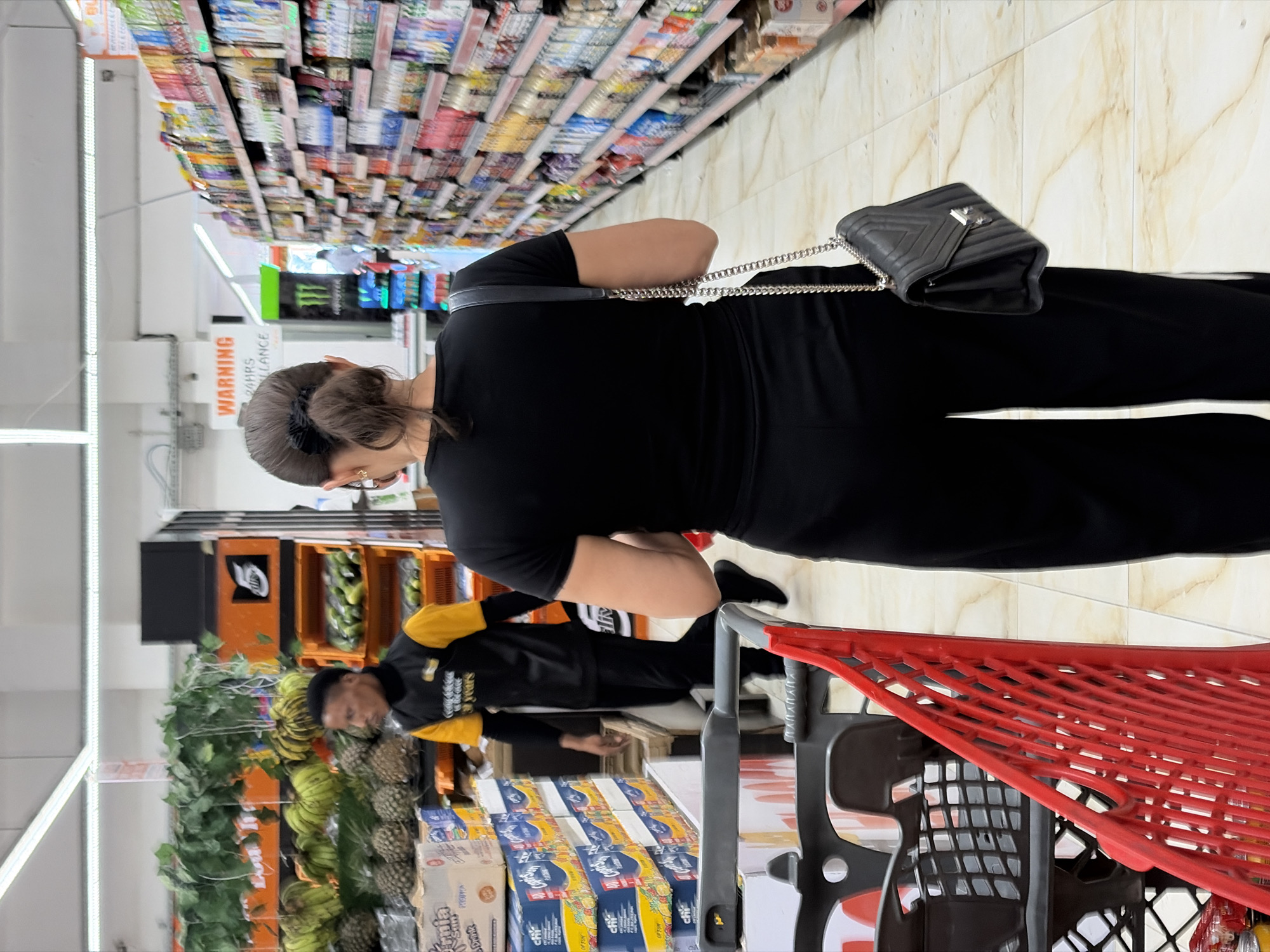 The image shows an indoor scene, likely a grocery store aisle in Igboefon, Nigeria. Shelves stocked with goods are visible on the left side of the image, receding into the background. A woman with her hair tied back is wearing black clothing and carrying a black purse with a chain strap. She is standing near a red shopping cart. A man wearing a black jacket with yellow sleeves is standing near a display of fruits, including bananas and pineapples. He appears to be stocking the shelves or arranging the produce. Boxes of juice are visible on a shelf near the shopping cart. The floor has a light, patterned tile surface.