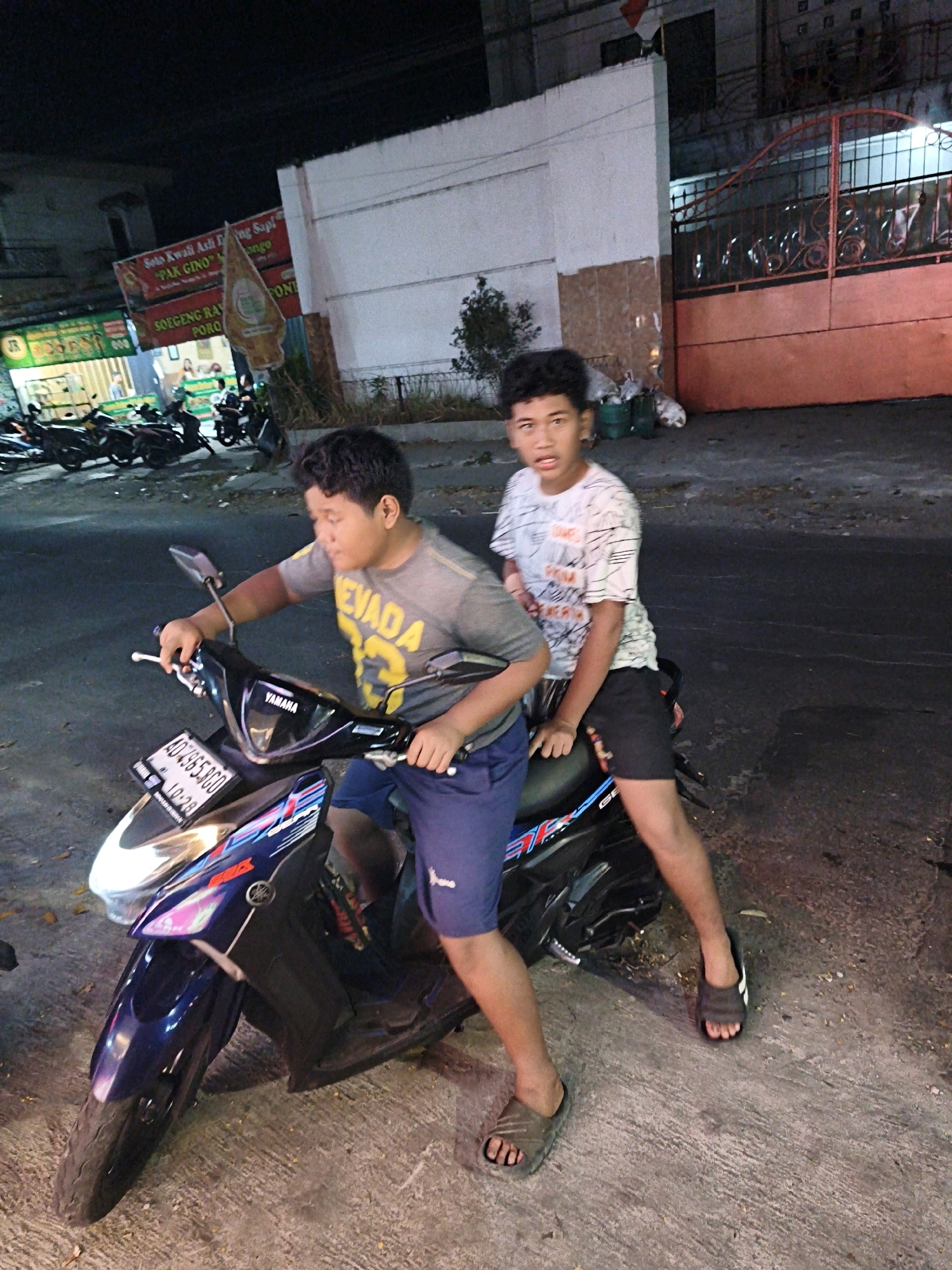 Two young males are positioned on and next to a dark blue Yamaha scooter parked on a paved surface. The male in the foreground, wearing a grey t-shirt with yellow lettering and blue shorts, is seated on the scooter, gripping the handlebars. The second male, wearing a white t-shirt with a black graphic print and black shorts, is seated behind him on the scooter. Both are looking towards the camera. The scooter has a license plate displaying "AD 4955 RGO". In the background, a building with white walls and a reddish-orange metal gate is visible, along with several parked motorcycles and a red and white flag. A sign with Indonesian text, possibly for a food establishment, is also present. The scene appears to be a street in the city of Surakarta, Indonesia, at night.