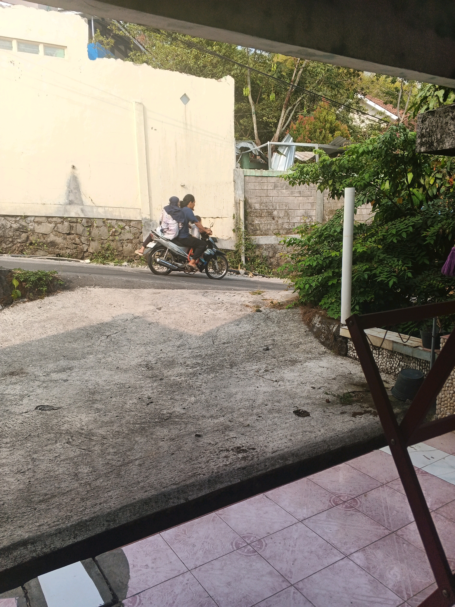 In Keji, Indonesia, two individuals are seen riding a motorcycle. The person in the front appears to be a man wearing a dark blue shirt, while the passenger behind him is a woman wearing a headscarf and a patterned top. The motorcycle is traveling on a paved road. To the left of the road, a light yellow wall with stone accents forms the boundary of a property. Beyond the wall, trees and vegetation are visible, suggesting a semi-urban or suburban setting. The foreground of the image shows a concrete surface and tiled flooring, indicating a pedestrian area or a viewpoint from a building. The overall scene depicts a moment of daily life on a street in Keji.
