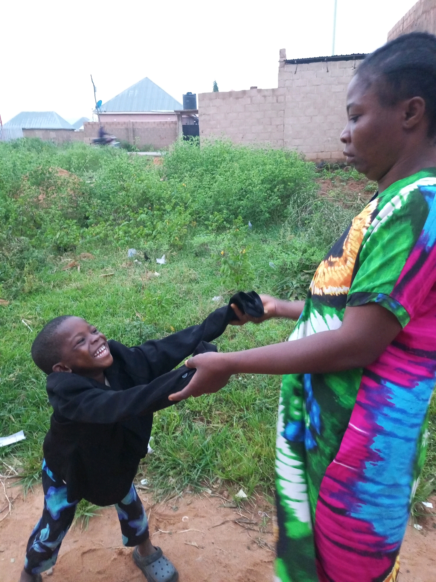 In Tempure, Nigeria, an adult woman wearing a colorful tie-dye dress with green, blue, pink, and yellow patterns is holding hands and playing with a young boy. The boy is wearing a black hooded jacket and patterned leggings with blue and black designs. He is smiling broadly and appears to be enjoying the interaction. They are standing on a dirt path, surrounded by green vegetation and some low-lying structures in the background, indicative of a residential area. The lighting suggests it is daytime, with overcast skies.