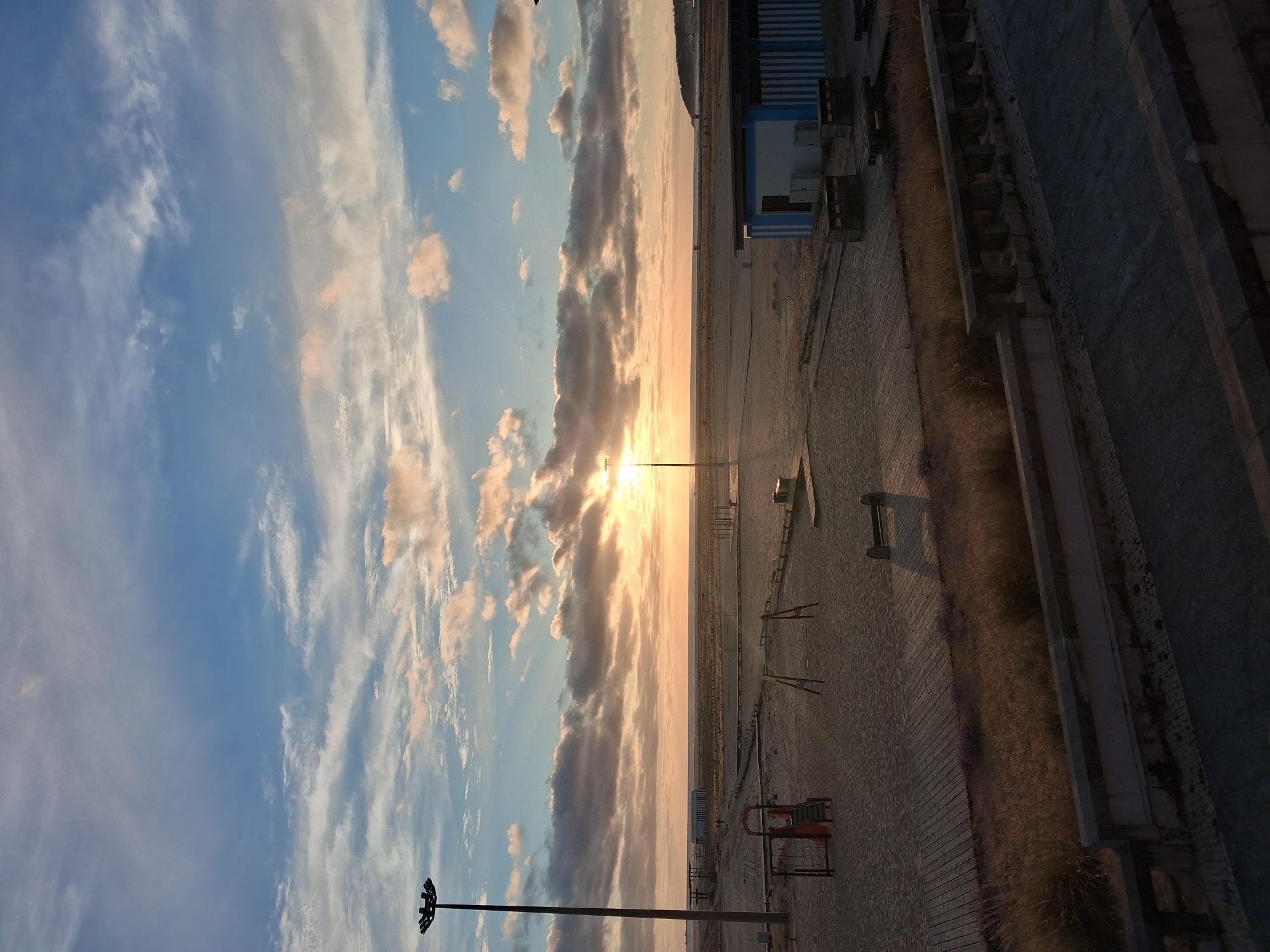 The image shows a beach scene in Figueira da Foz, Portugal, during sunset. The sky is a gradient of blue to orange, with clouds lit by the setting sun. The beach appears to be sandy, with a few structures, possibly beach huts or facilities, and a promenade running alongside it. Benches are visible on the beach. The image is taken from an elevated perspective, giving a broad view of the beach and the coastline. A light post is in the foreground. The sea is a thin horizontal line between the beach and the sky.