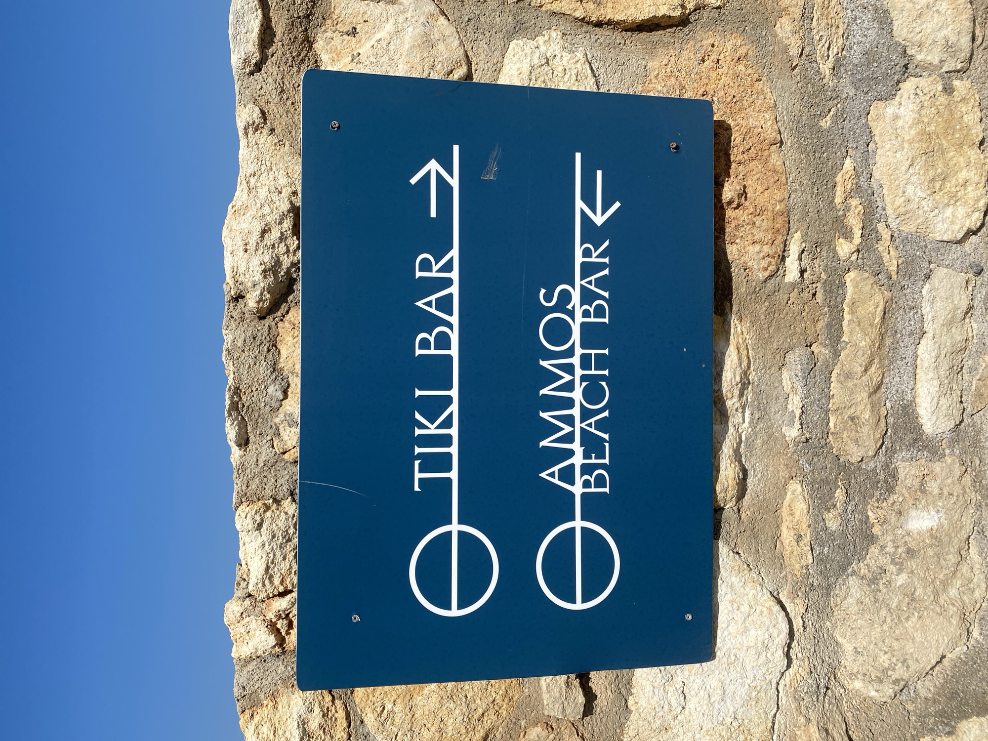 A blue sign with white text is attached to a rough stone wall under a clear blue sky. The sign has two directional indicators. The top line reads "TIKI BAR" with an arrow pointing to the right. The bottom line reads "AMMOS BEACH BAR" with an arrow pointing to the left. The sign is mounted on a wall made of irregular, beige and gray stones. The location is Panormos, Greece.