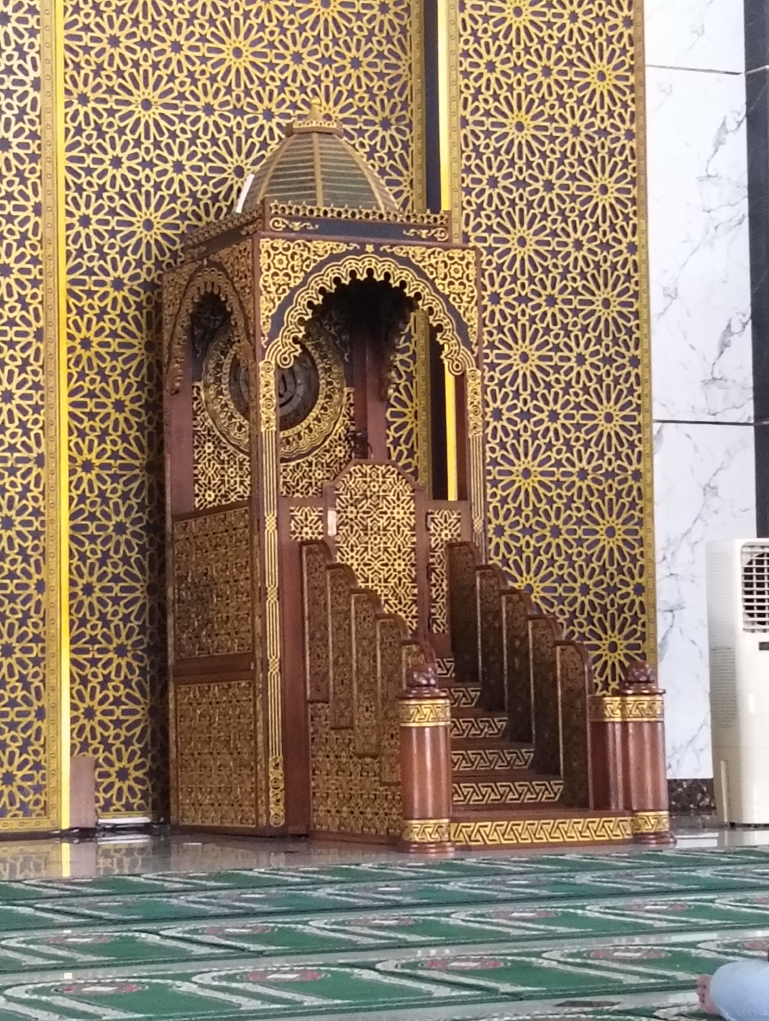 The image depicts the interior of a mosque, specifically showing a minbar (pulpit) adorned with intricate golden geometric patterns on a deep purple background. The minbar is made of dark wood with elaborate carvings and a tiered structure leading up to its top. The surrounding walls also feature the same repeating golden geometric design. To the right of the minbar, a portion of a white air conditioning unit is visible, and further right, a marbled wall section. In the foreground, green prayer mats are laid out on a polished floor, indicating the space is prepared for prayer. The scene appears to be within a mosque in Surabaya, Indonesia, given the architectural style and the presence of prayer mats. No people are clearly visible in the frame, except for a partial view of a person's legs in the bottom right corner. The lighting is moderate, illuminating the decorative elements of the minbar and the patterned walls.