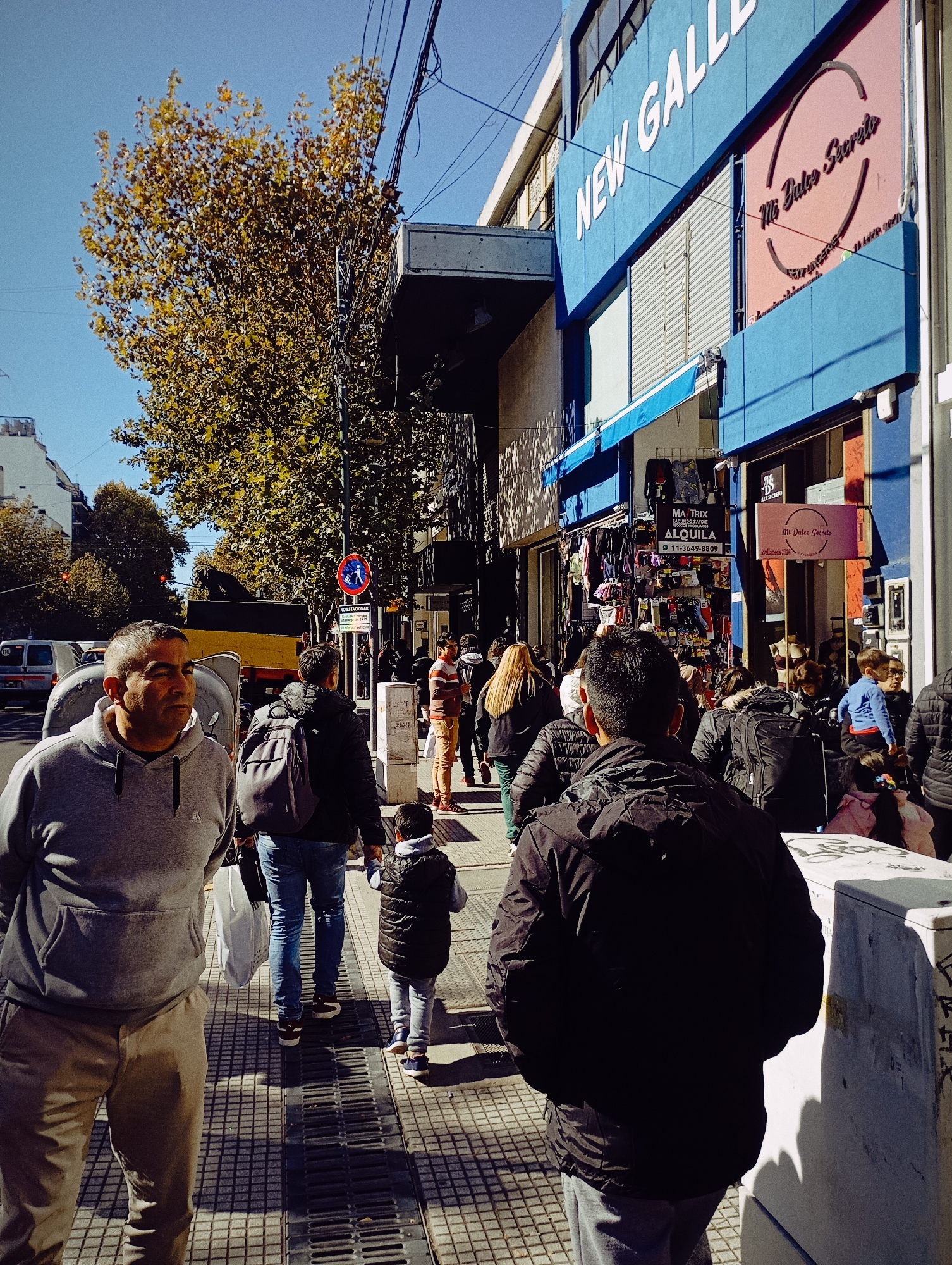 A sunny day on a busy street in Buenos Aires, Argentina. Pedestrians are walking along a tiled sidewalk. Buildings line the street, one with a blue facade and the words "NEW GALLERY" in white. Signs for "Mi Dulce Secreto" and "Matrix Alquila" are visible. Trees with autumn foliage are present. A yellow truck is partially visible in the street. The pedestrian traffic sign reads "NO ESTACIONAR".