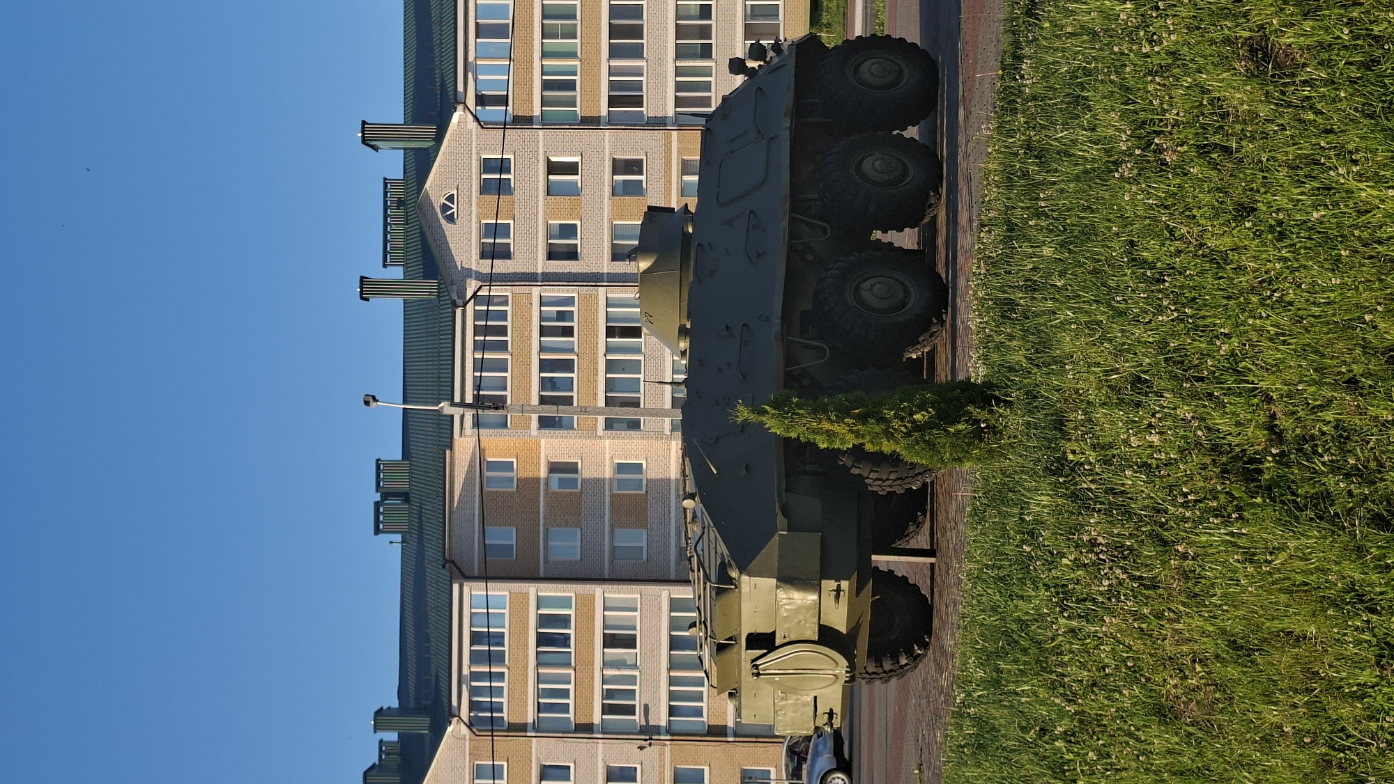 An olive-drab, eight-wheeled armored personnel carrier, possibly a BTR-80 variant, is parked on a paved surface in front of a modern, multi-story apartment building with beige and light grey brickwork and a dark green tiled roof. The building has numerous windows with white frames. The APC has a turret and is positioned facing left. A small, conical evergreen tree stands in front of the APC. The foreground is a grassy area with small white clover-like flowers. The sky is a clear, bright blue. The scene appears to be in a residential area, consistent with the city of Smolevichi, Belarus, given the architecture and common use of such vehicles as monuments or displays in Eastern Europe. There are no people visible in the frame.