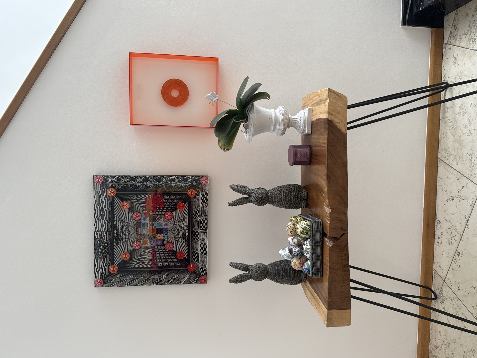 A vertically-oriented photograph captures a wall with several decorative elements. Starting from the top left, there's an orange square frame with a circular design inside. Below it, a framed piece of art is displayed, featuring intricate geometric patterns in black, white, and various colors. Moving towards the right, an orchid in a white, ornate vase is attached to a wooden shelf. The shelf also holds a purple candle jar, two woven bunny figures, and a woven basket filled with decorative eggs. The wooden shelf is supported by black hairpin legs. The wall is painted white. The photograph does not provide sufficient information to verify the location in the city of San Bernardino Tlaxcalancingo, Mexico.