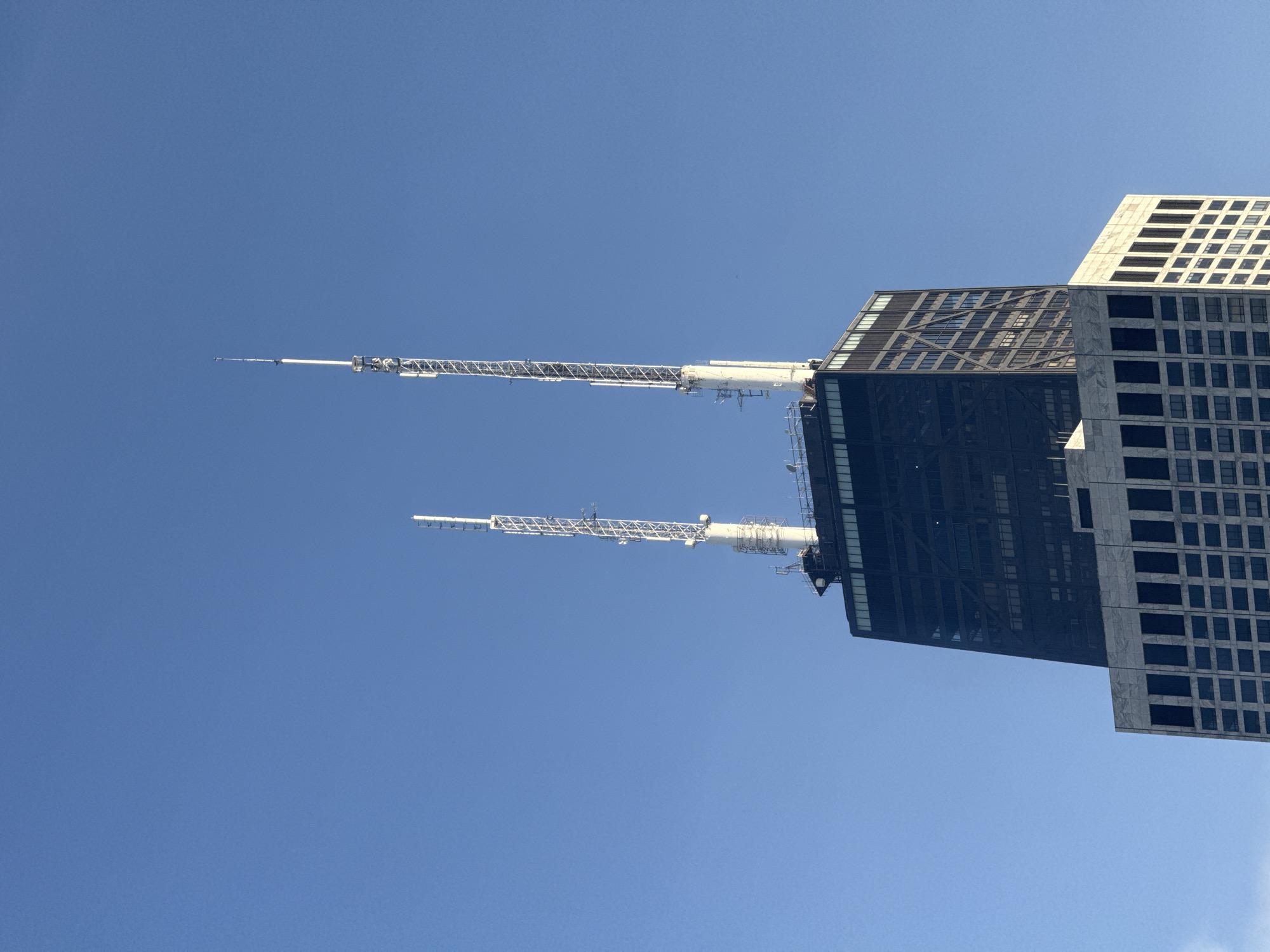The image shows a close-up view of the top portion of the John Hancock Center, a skyscraper located in Chicago, Illinois, United States of America. The building features a distinctive dark, X-braced structure at its upper levels, topped by two prominent white antennas. The lower section of the visible building has a lighter, grid-like facade. The sky behind the building is a clear, vibrant blue, suggesting fair weather. No people or other objects are visible in the frame.