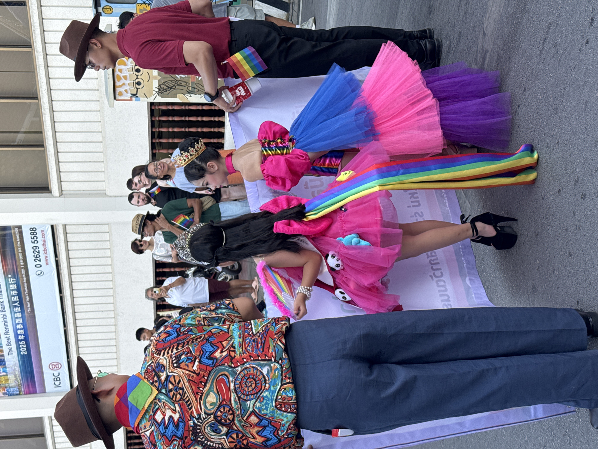 The image shows a scene at a Pride parade in Chiang Mai City Municipality, Thailand. In the foreground, a person wearing a colorful patterned shirt, blue pants, and a brown hat is partially visible. In front of them, two individuals are dressed in bright, vibrant costumes. One wears a pink dress adorned with rainbow accents, including a large rainbow sash and a rainbow fan. They also wear a crown and high heels. The second person is dressed in a similar fashion with a colorful dress in the colors pink, blue, and purple, also with a crown. Behind them, a group of people, some wearing Pride-themed clothing and accessories, are partially visible. A man in a red shirt and black pants, also holding a small rainbow flag, is seen above the person in the colorful dress. A banner is visible beneath the individuals in costume. In the background, a building with the logo of the Industrial and Commercial Bank of China (ICBC) and a phone number are visible.