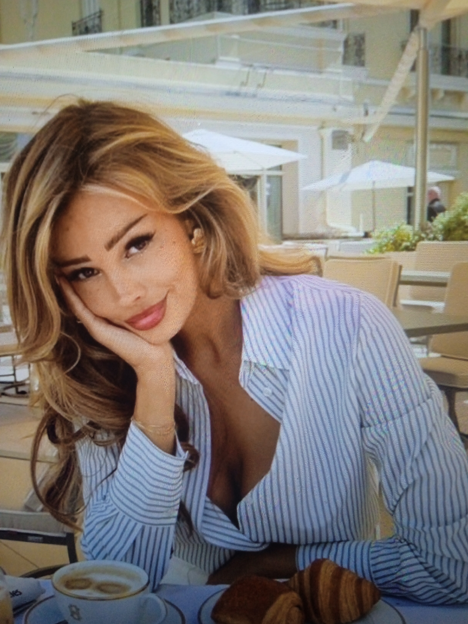 A woman with blonde, wavy hair is shown from the chest up, resting her left cheek on her hand. She is wearing a blue and white striped button-down shirt, unbuttoned to reveal cleavage. Her right arm is bent, with her hand resting near her chest. In the foreground, a cup of coffee with foam art and two croissants are on a table. The background appears to be an outdoor cafe with tables and umbrellas, suggesting a pleasant day. The image is a photograph, likely taken outdoors. There is no information within the image or its context to suggest a location in the city of Chernivtsi, Ukraine.