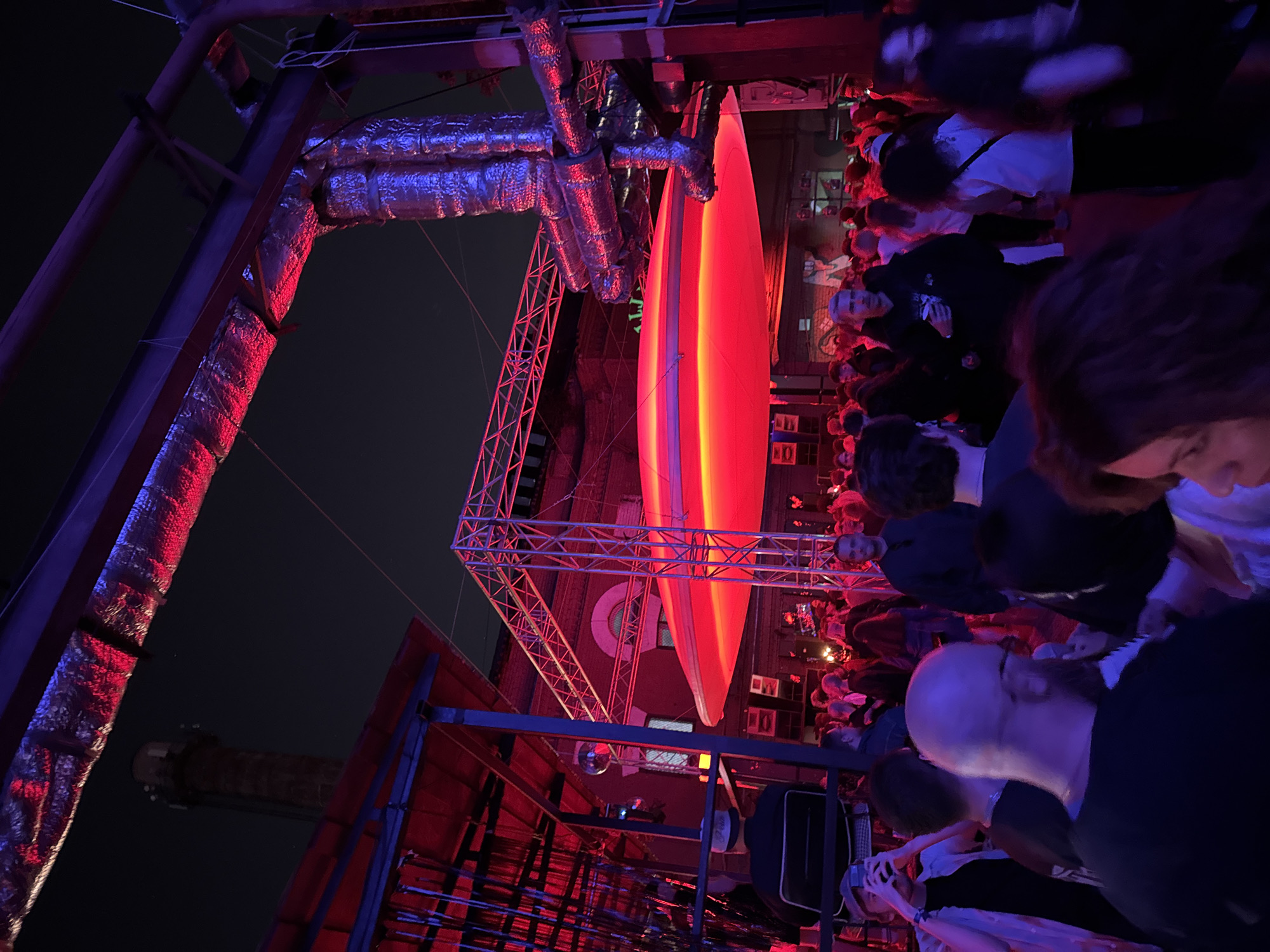 The image captures a nighttime event at Mutabor, a cultural and event space located in a repurposed industrial complex in Moscow, Russia. The scene is illuminated by intense artificial red and blue lighting.
A large gathering of people, mostly adults, occupies the central and foreground areas of the image, standing and moving within what appears to be an outdoor or semi-enclosed industrial courtyard. Overhead, a large, flat, circular or oval-shaped light fixture glows a vibrant red, suspended from a metal truss framework. Surrounding and above the crowd are various industrial structures: exposed metal beams, additional truss scaffolding, and insulated ventilation ducts wrapped in reflective silver material, all reflecting the red and blue light.
In the background, against a dark night sky, a tall, slender brick industrial chimney with communication equipment at its top is visible on the far left. Adjacent to it, older brick buildings with distinct arched windows are partially visible, consistent with a former factory or plant. A small disco ball is suspended on the left side of the scene.
Among the crowd, individuals are observed standing, conversing, and engaging in activities typical of a social gathering. On the left side, a person holds a smartphone up, seemingly recording the event. Several individuals are holding drinks. Clothing styles are predominantly casual and dark, with details becoming apparent under the strong colored light.