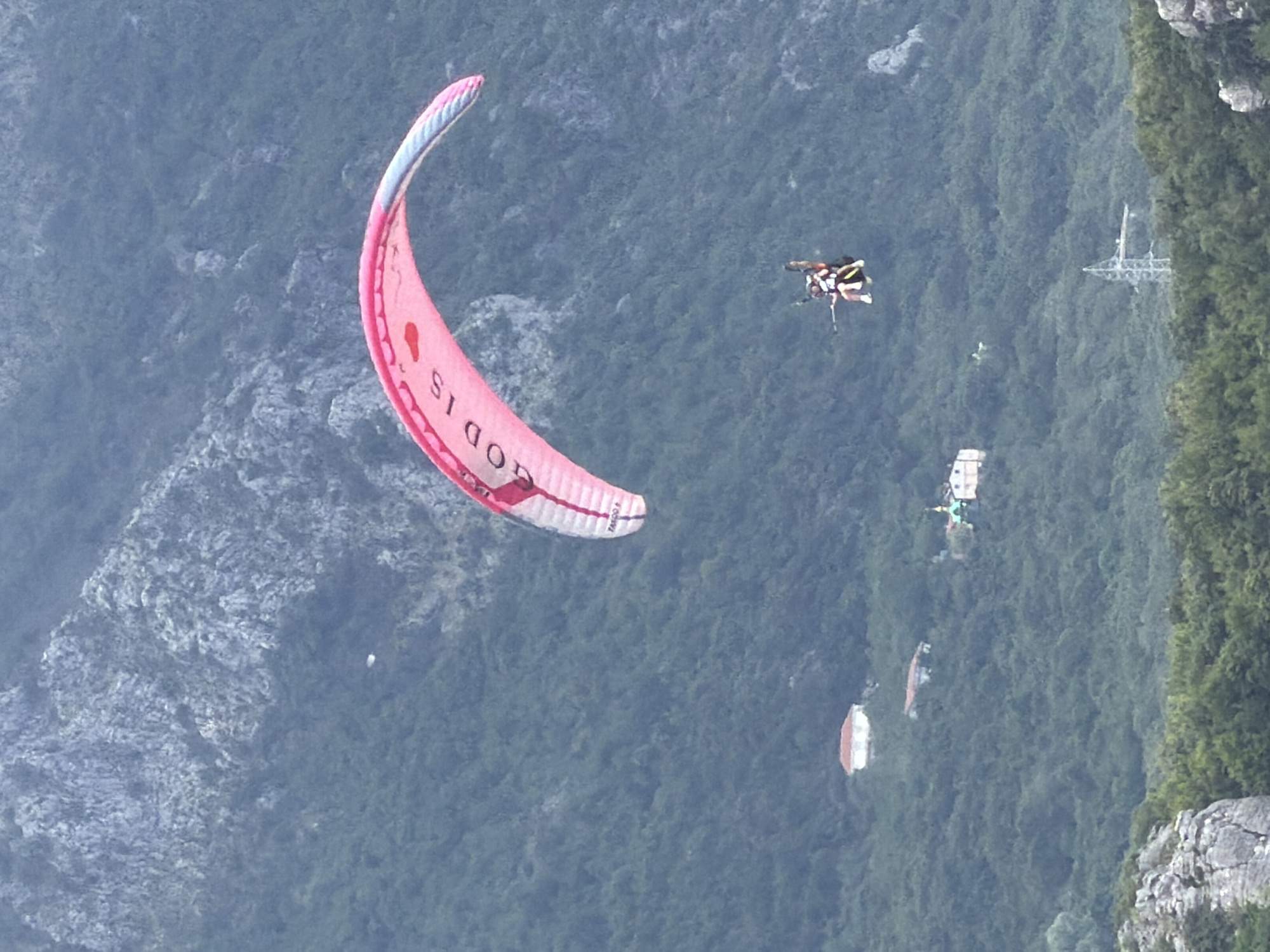 This image captures an aerial view over a lush, mountainous landscape in Boreti, Montenegro. The scene is dominated by a large, curving pink and white paraglider in the foreground, with visible text "GOD IS" and partially "TANGO P" on its canopy.
Below and to the right of the large paraglider, a tandem paragliding pair is visible, suspended in harnesses, indicating an active paragliding event or flight. A small, striped, insect-like object, resembling a wasp or bee, is also visible in the mid-ground, suggesting it's close to the camera or airborne.
The background reveals dense green forests covering steep slopes and rocky outcrops. Scattered across the landscape are several structures, including a distinctive power line tower and various buildings with red and white roofs. Another object on the ground, possibly a discarded or landed paraglider canopy, can be seen.
The lighting suggests daytime, possibly under overcast or hazy conditions, lending a somewhat muted tone to the colors of the landscape. The image is oriented as if looking directly downwards, with the paragliders flying across the frame.