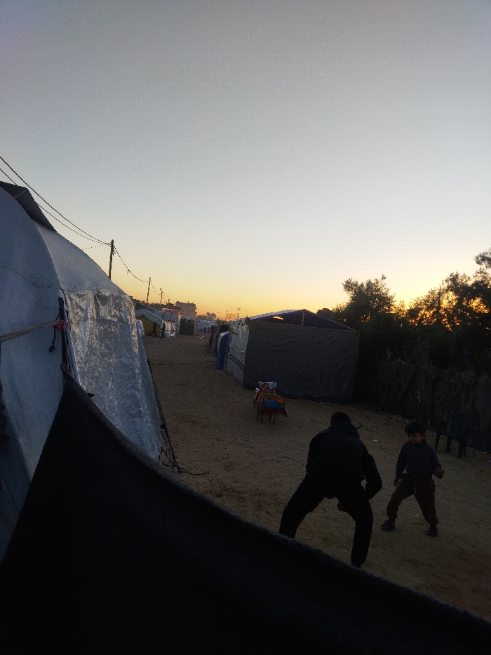 The image depicts an outdoor scene at dusk in Khan Yunis, Palestinian Territory. The sky displays a gradient from light grey overhead to an orange-yellow glow at the horizon, indicating sunset. A sandy or dirt path extends from the foreground into the distance, flanked by rows of makeshift shelters and tents constructed from various materials including tarpaulins (light blue, silver, dark green). On the left, a large, light-colored tent occupies the foreground, secured with visible blue and red ties. Overhead, utility poles support power lines running parallel to the path. On the right, a dark green tent is visible, with a small, multi-colored stool positioned near its entrance. Further right, dark foliage from trees and bushes is silhouetted against the setting sun, where two dark plastic chairs are also present. In the mid-ground, two figures are visible: an adult in dark clothing is crouched or bending forward, facing right, and a child in a dark top and lighter trousers stands to the adult's right, facing towards them.
