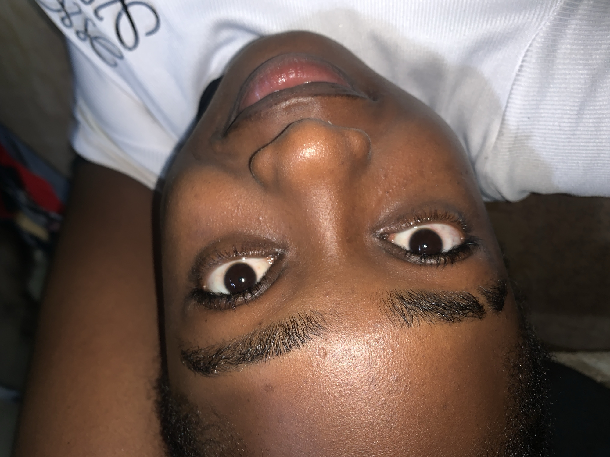 The image shows a close-up, overhead view of a person's face lying on their back. The person has dark skin, dark eyebrows, and dark eyelashes. Their eyes are open, and their mouth is slightly parted, revealing their tongue. The person is wearing a white t-shirt with a black design on it. The background is blurred, but it appears to be a room. The image is not geographically specific and does not provide any information about the city of Rafin Par, Nigeria.