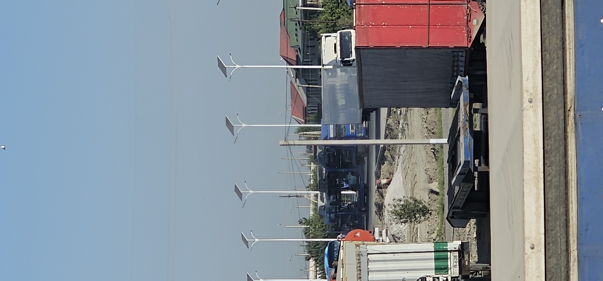 The image depicts a street scene in Sheyhancha, Uzbekistan, on a clear day with a pale blue sky. In the foreground, a large blue flatbed trailer is visible. Behind it, to the left, is the side of a white and green truck, with a license plate reading "R-628 T". To the right of the trailer is a large red trailer. In the middle ground, a blue car carrier truck is transporting multiple vehicles. To its right, a white truck is parked. Further back, several houses with red and green roofs are visible, along with numerous streetlights equipped with solar panels. The overall environment appears to be an open area, possibly a staging or parking lot, with some construction or excavation work evident. There are no people clearly visible in the image.