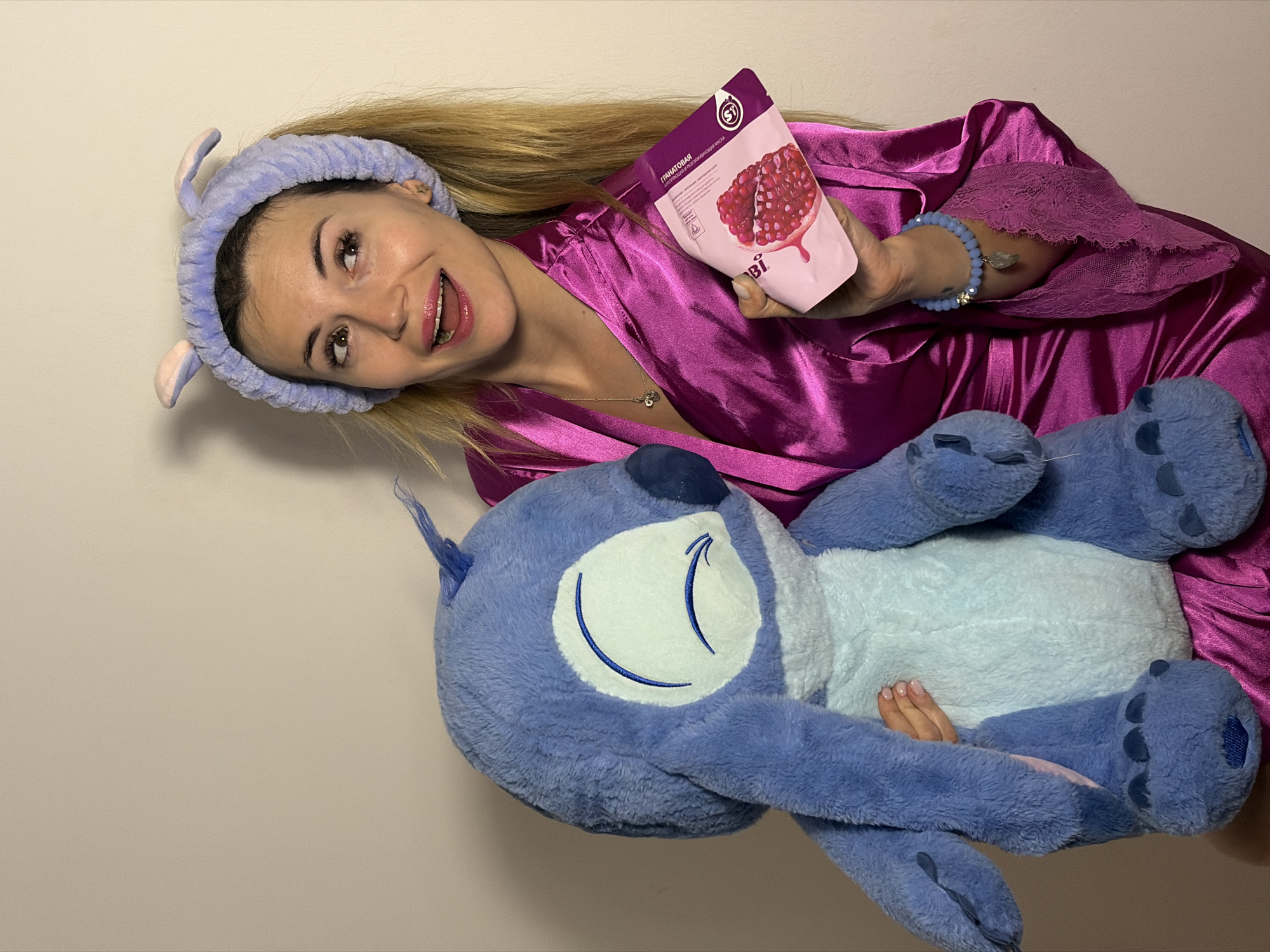 A woman wearing a lavender headband with small bunny ears, and a shiny purple silk robe, holds a pink facial mask pack with a pomegranate depicted on it. She is lying down and hugging a blue and white Stitch plush toy. She wears a bracelet on her right wrist and a necklace. The background is a plain light beige wall. The location is not specified in the image.
