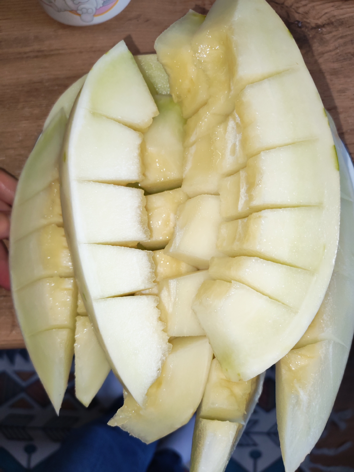A close-up shot shows a platter of cut honeydew melon. The melon is sliced into bite-sized pieces and arranged on a plate. The plate appears to be white with a blue rim. The melon is pale green with yellow flesh and is glistening with juice. The background shows a wooden surface and a colorful patterned floor. The image was taken indoors. The location in Izhevsk, Russia cannot be determined from the visual information provided.
