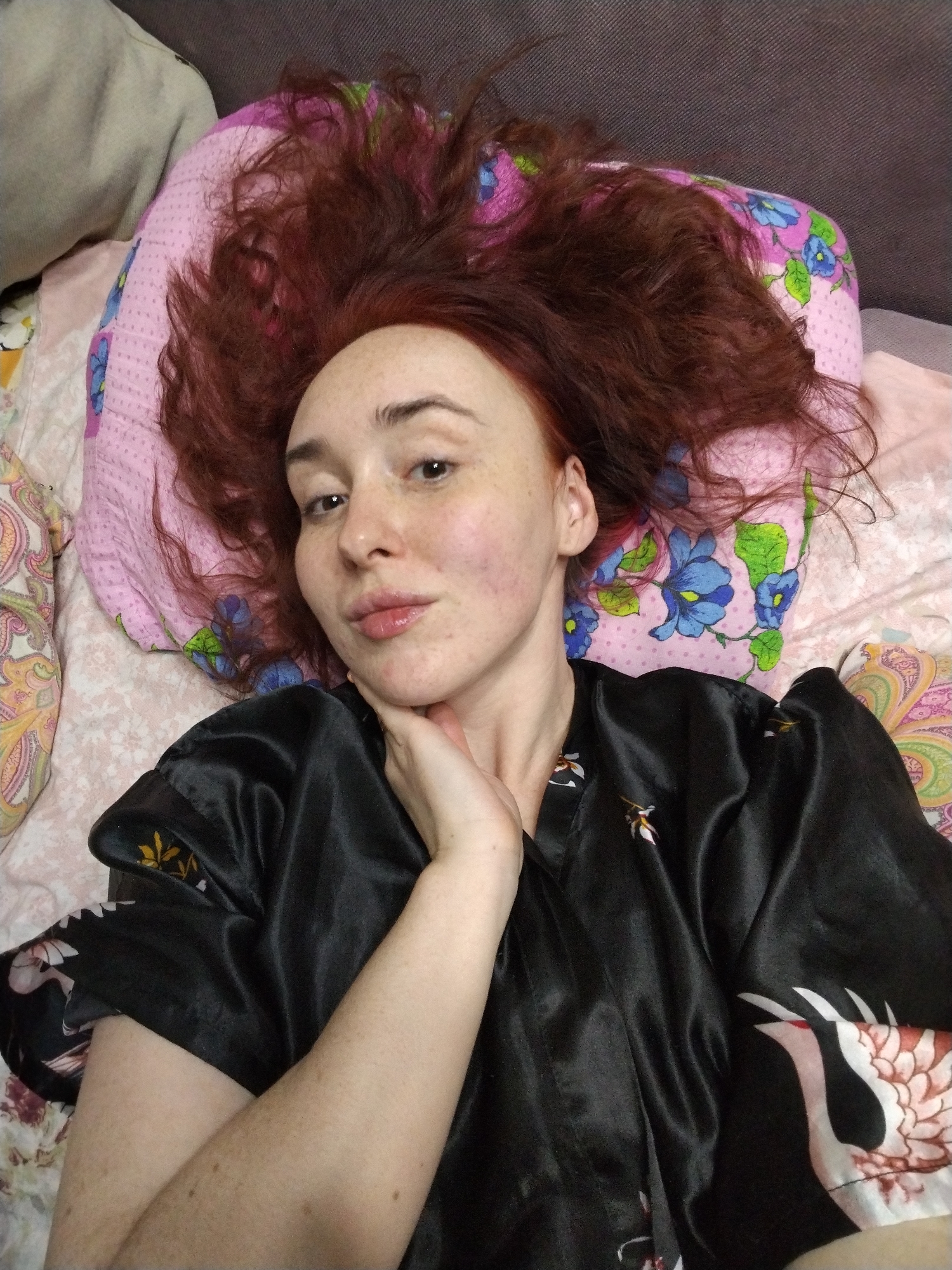 The scene depicts a female individual lying supine, viewed from a high angle. Her reddish-brown, wavy hair is spread across a pink pillow patterned with blue flowers and green leaves. The individual has fair skin with visible freckles on her face and arms. Her eyes are open and directed at the viewer, and her lips appear glossy. Her right hand is positioned beneath her jawline. She is wearing a black, satin-like robe featuring a white and red crane or bird design on the right side and yellow/gold leaf motifs. The surrounding environment includes light-colored bedding with subtle patterns, a multi-colored paisley patterned pillow to the left, and a dark grey textured surface in the background. The precise location within Izhevsk, Russia, is not discernible from the visual evidence provided.