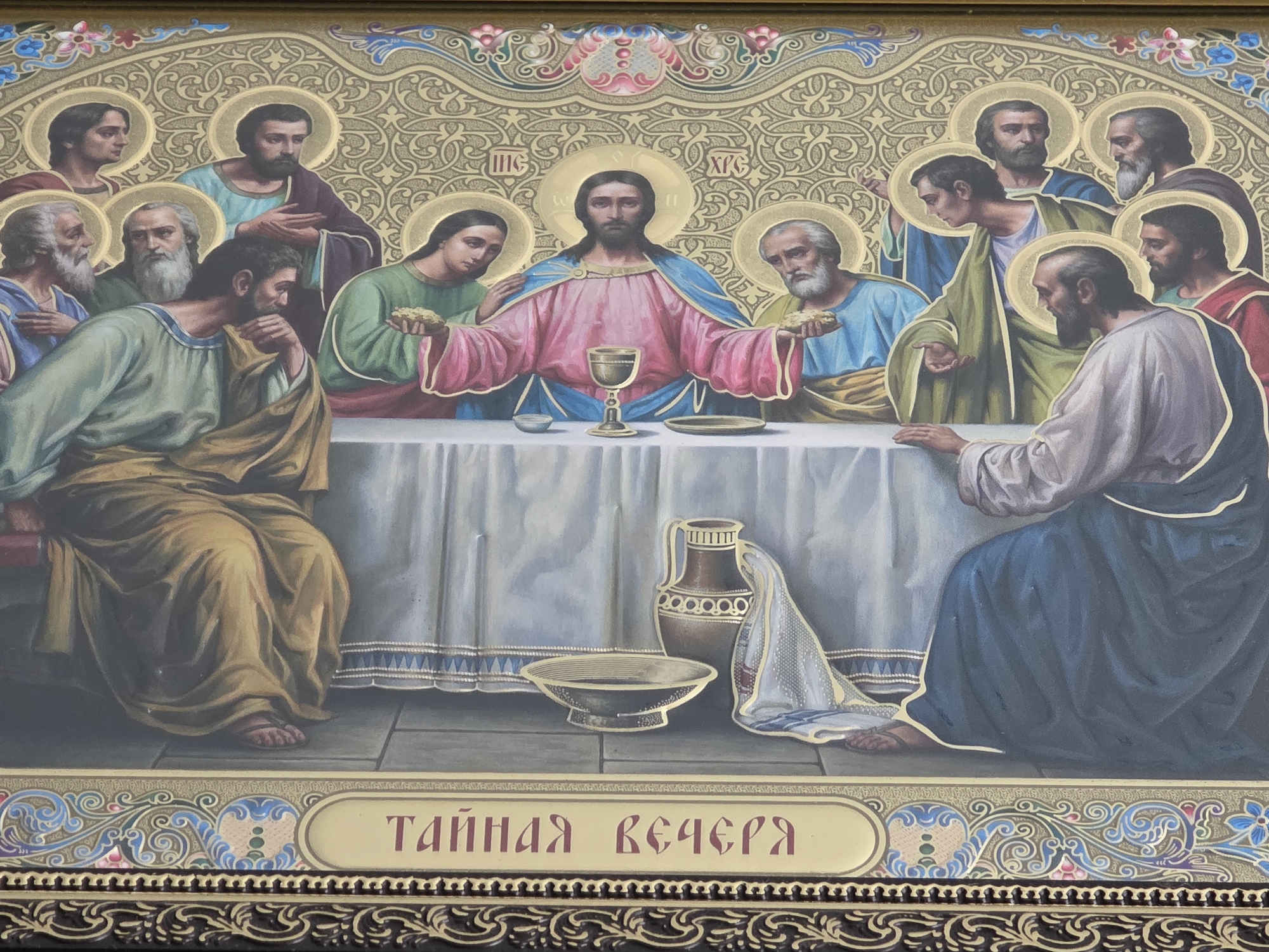 This is a close-up, eye-level, horizontal shot of an icon depicting the Last Supper. The scene is set around a long table draped with a white tablecloth. At the center of the table, Jesus, identified by his halo and pink and blue robes, holds a chalice. He is flanked by his disciples, who are also depicted with halos. Some disciples are reaching for food, while others are looking towards Jesus. The disciples are dressed in robes of various colors, including green, blue, gold, and red.
The background of the icon is ornate gold with intricate patterns. Below the table, there are objects such as a brown jug, a bowl, and a folded white cloth. The floor appears to be tiled. The overall mood of the painting is somber and reverent, consistent with a religious depiction.
At the bottom of the icon, there is a decorative border with floral and swirling patterns. Centered within this border, on a cream-colored background, is text in red Cyrillic letters: "ТАЙНАЯ ВЕЧЕРЯ". This translates to "The Last Supper." The entire image is framed by a darker border, likely part of the icon's casing. The lighting is even, suggesting an indoor setting, and there are no indications of time of day or weather.
