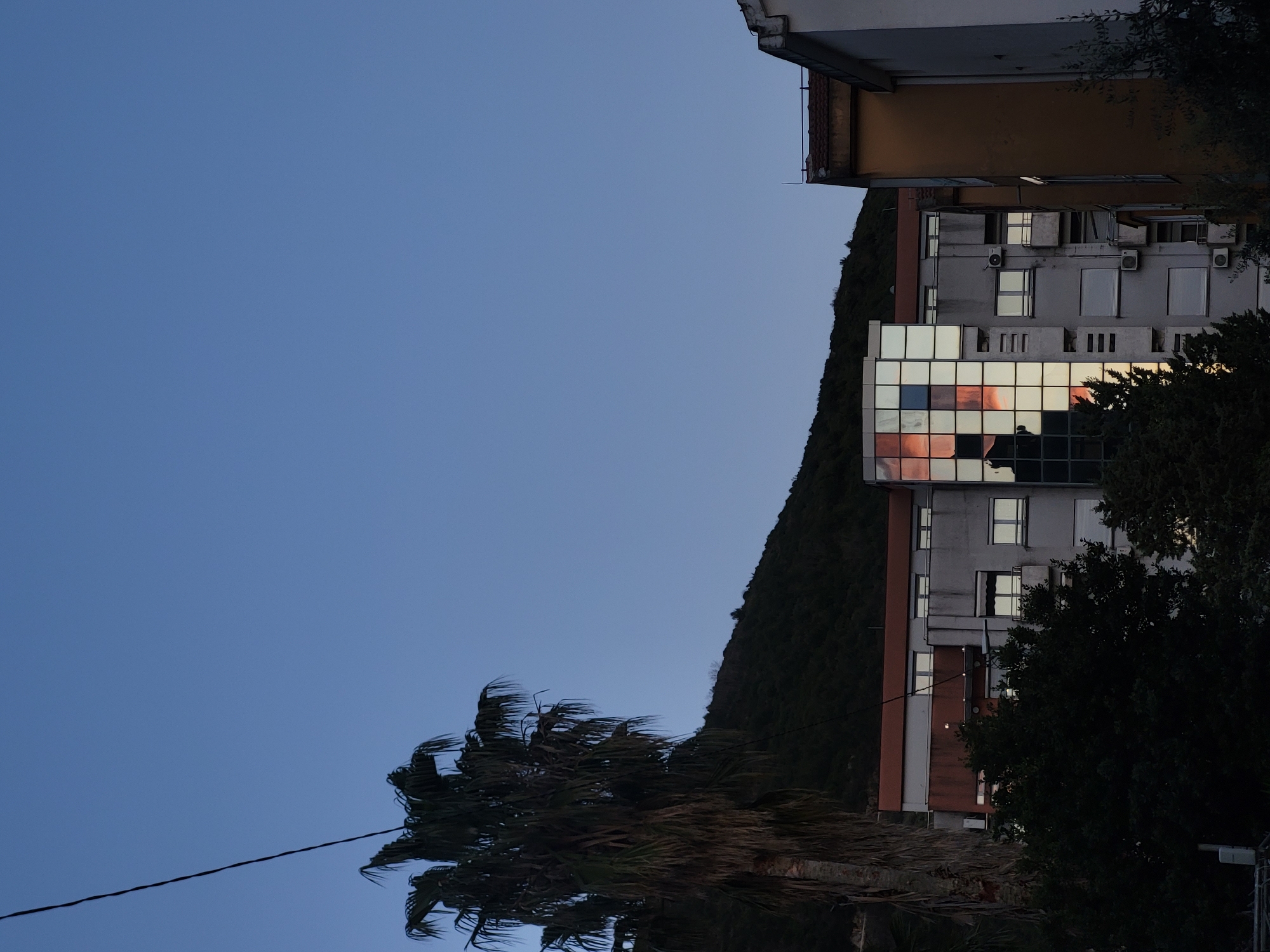 The image shows a building in Budva, Montenegro, with a mirrored facade reflecting the sunset. A tree is in the foreground, and a mountain is in the background. The sky is blue. A power line is visible in the lower left corner.