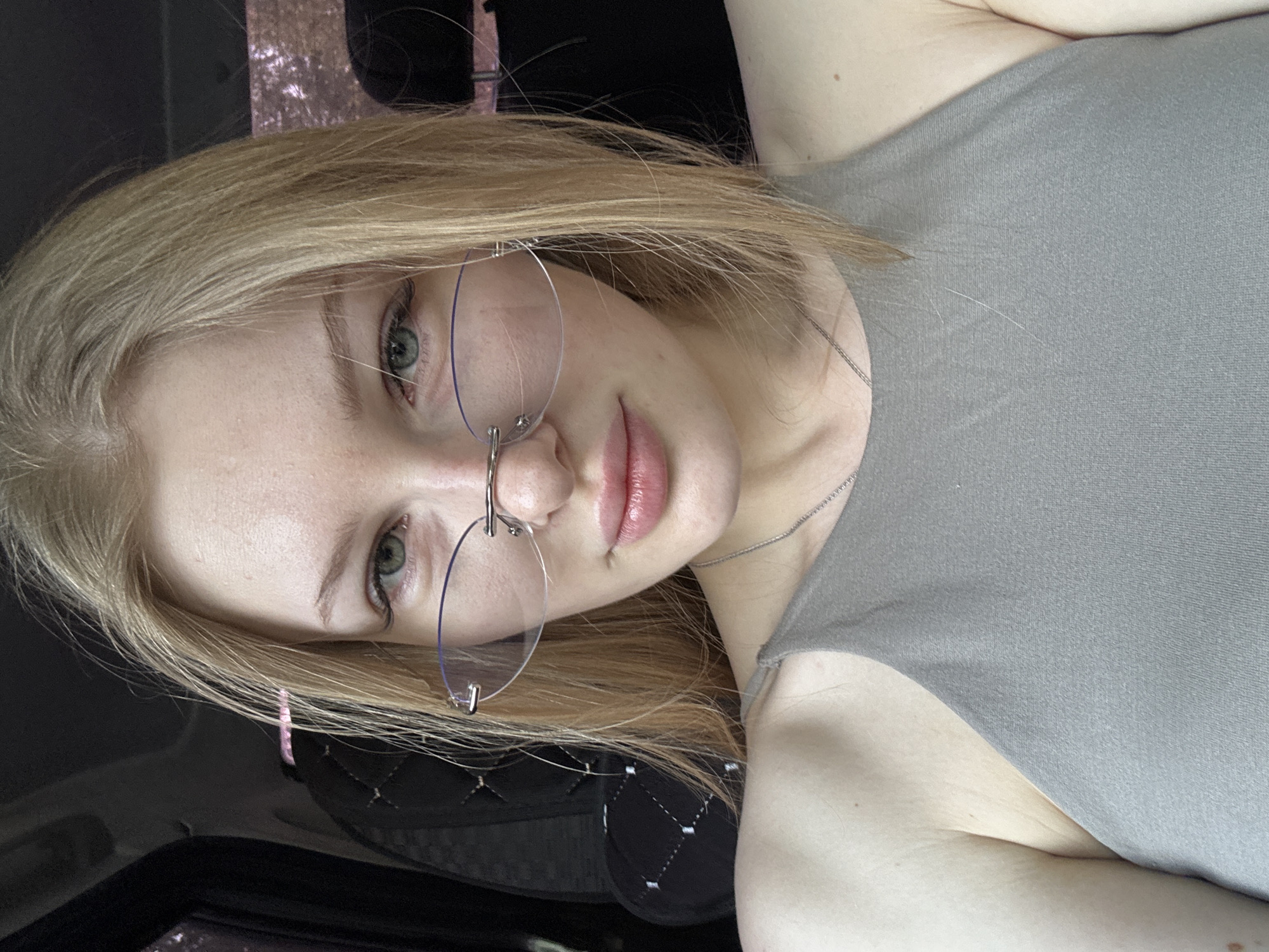 A photograph depicts a fair-skinned young woman with blonde, shoulder-length hair and green eyes. She is wearing rimless glasses and a grey sleeveless top. The photograph is taken from an unusual angle, with the woman's head tilted to the left. The background appears to be the interior of a vehicle, showing parts of a seat and a dashboard. There is no visual information to suggest the location is Perm, Russia.