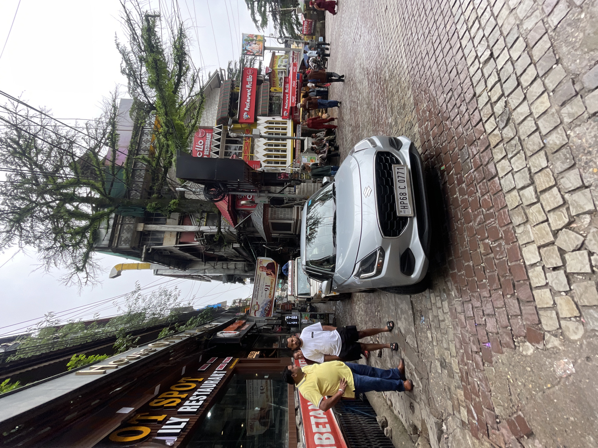 The image depicts a street scene in Bhagsu, India, likely on a cloudy day. In the foreground, a silver Suzuki car is parked on a wet, cobblestone street. The car's license plate reads "HP68 C 0771". To the left of the car, two men are standing. The man on the left wears a yellow polo shirt, blue jeans, and brown shoes. The man on the right wears a white t-shirt, black shorts, and black sandals. Behind them, a building with storefronts lines the street, featuring signage for "OT SPOT ILY RESTAURANT". Further down the street, other businesses and buildings are visible, including "Restaurant McLo" and signs with Hindi text. Several people are walking and gathered on the sidewalk, and a few vehicles, including what appears to be a white jeep, are parked further down the street. Trees with mossy branches are visible in front of some buildings, and electrical wires crisscross overhead. The overall impression is a bustling, narrow street lined with shops and businesses in a hilly region.