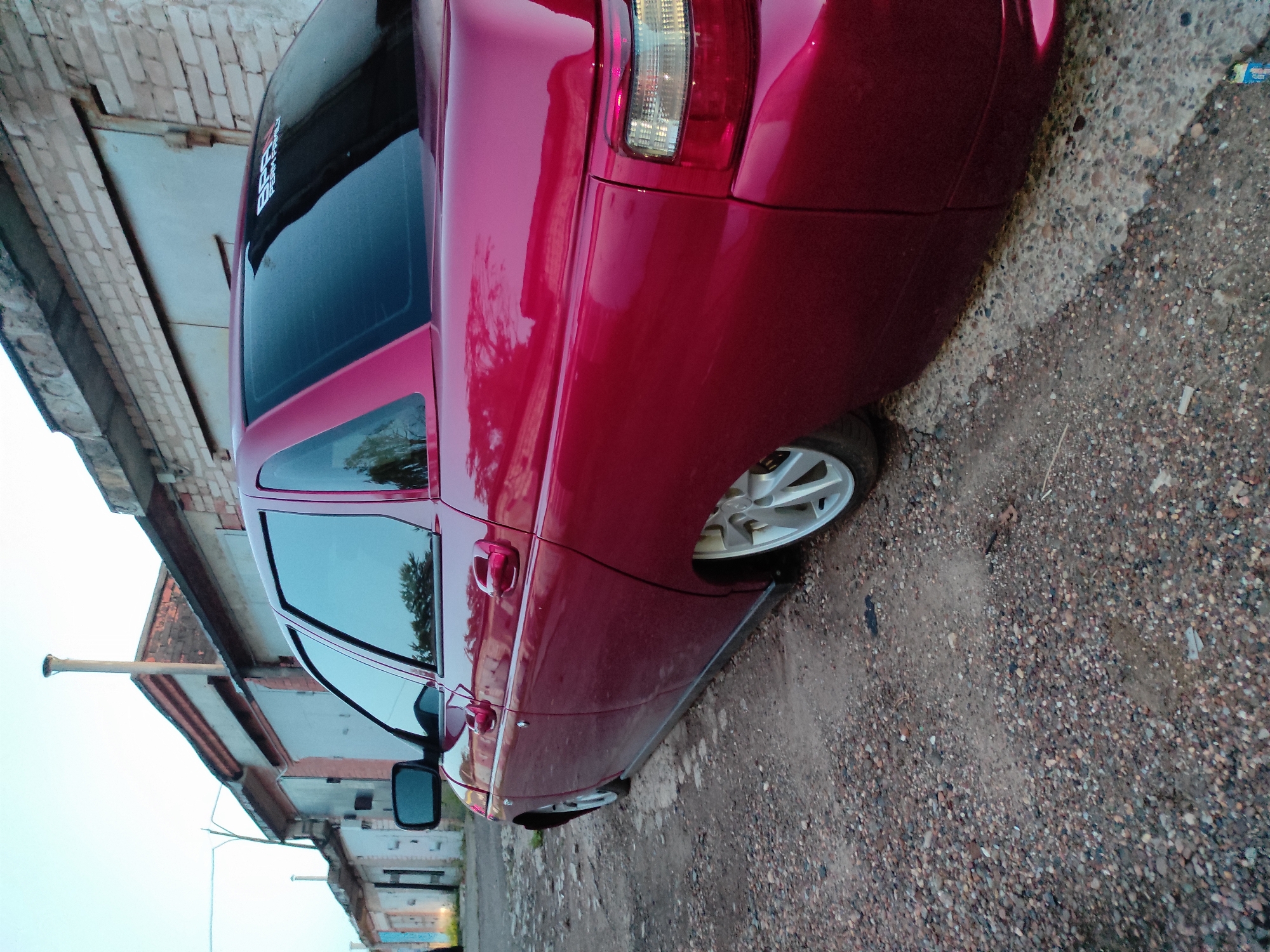 A red four-door sedan, possibly a Lada model, is parked on a gravel and dirt path next to a row of old brick buildings. The car is the primary subject, shown from its rear-left side, and appears to be heavily customized with white multi-spoke alloy wheels and tinted windows. On the upper part of the rear window, a white sticker with stylized text is visible, prominently featuring the letters "BB" and additional, less discernible text below it.
The scene depicts an urban or semi-industrial environment, possibly an alley or a rear service area. The buildings, constructed from brick, show signs of age and wear, consistent with older garages or industrial structures. The ground is an unpaved mix of small stones, gravel, and dirt. A thin pole, potentially a streetlight, is visible in the background on the left.
The time of day appears to be either early morning, late afternoon, or an overcast day, as indicated by the soft, diffused lighting and bright, cloud-covered sky. The weather seems calm. No people or activity are present, suggesting the car is stationary and unoccupied. The overall setting and car model are consistent with a location like Chaikovsky, Russia.