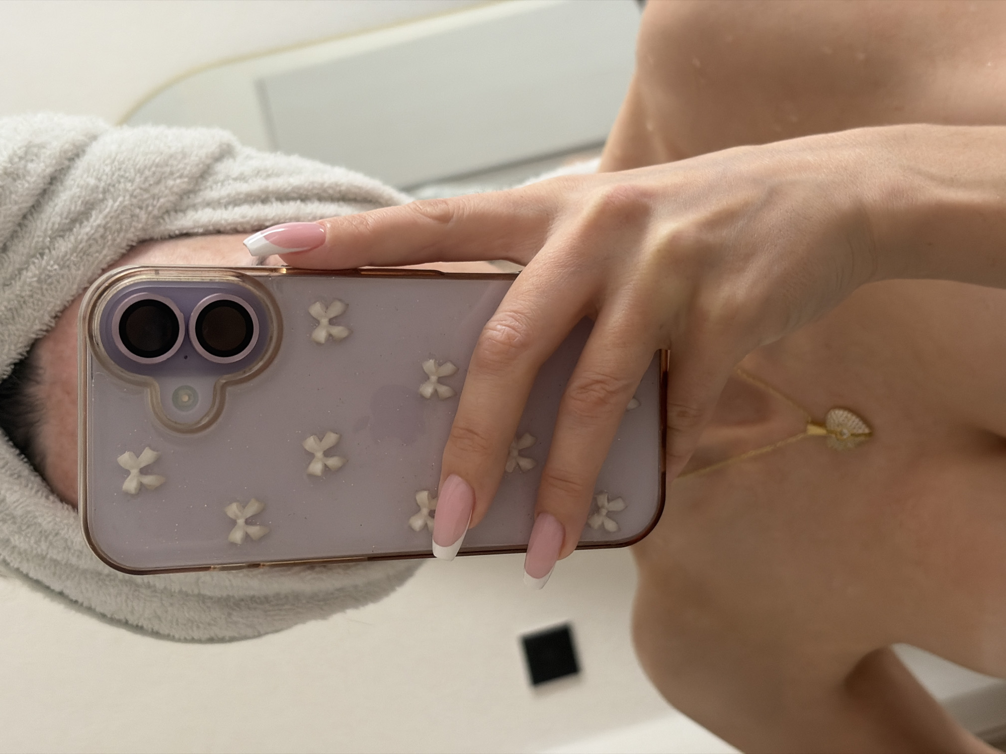 A person is captured in a mirror selfie, primarily showing their upper body from the chest up. Their head is wrapped in a textured, light grey towel, suggesting they have recently showered or bathed. The individual holds a smartphone with their right hand, positioning it to take the photo.
The smartphone features a clear or translucent light purple case adorned with several small, white, three-dimensional bow-shaped embellishments. The dual camera lenses of the phone are visible on the upper left side of the case. The person's nails are manicured with a classic French tip design, having a pink base and crisp white tips.
A delicate gold chain necklace with a shimmering, shell-shaped pendant hangs around the person's neck. Their bare shoulder and upper torso are visible, showing smooth skin that appears slightly damp or glistening. The background is generally bright and out of focus, consisting of light-colored walls or surfaces, consistent with an indoor setting like a bathroom. A small, dark square shape is reflected on the surface below the phone. There is no visible text in the image. The scene depicts a casual, personal moment. The provided location context is Saint Petersburg, Russia.
