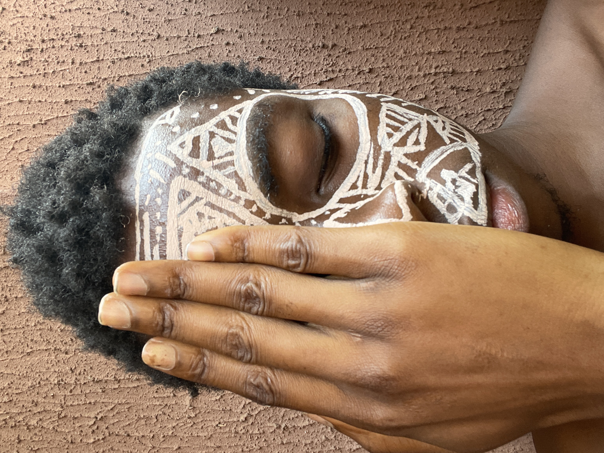 A person of African descent lies on their side against a textured brown surface resembling a wall. The person's head is turned toward the viewer, with a closed eye visible. The face is painted with white geometric designs, and the person's hand is positioned over their mouth. The head is surrounded by dark, curly hair. The scene appears to be indoors. There is no contextual information in the image to place it in Nkpor, Nigeria.