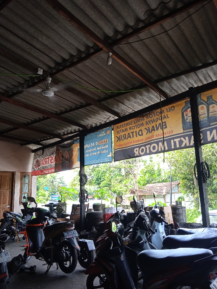 This image depicts the interior of an open-air motorcycle repair shop or garage, likely located in Patuk, Indonesia. The structure features a corrugated metal roof supported by wooden beams, with a ceiling fan actively rotating, suggesting a warm climate during the daytime. Natural light filters in from the open side, which looks out onto a lush green landscape of trees and foliage, with glimpses of a residential building with a tiled roof in the distance.

Numerous motorcycles, primarily scooters, are parked throughout the space, some appearing to be on stands, indicating ongoing or pending repair work. Black barrels are visible near the open entrance, and stacked tires can be seen in the background. A light-colored building with wooden doors and windows forms a partial back wall.

Several large banners are prominently displayed, hung from the ceiling beams. These banners, largely mirrored, advertise motorcycle-related products and services. Interpreted text includes phrases like "SHELL SINTETIK MOTOR OIL," "AYAMOYAL MOTOR HITAM," "ENAK DITARIK," and "HARGA BERSAHABAT." Other text visible includes "MOTOR SAKTI," "ASTRA," and "BARU 2.0 LITER." A small white sign reads "ELOIN MOTOR." On a motorcycle in the lower left, a license plate "AG 2956 RV" with "02-26" is visible. No people are explicitly visible in the frame, but the active fan and arrangement of motorcycles suggest a working environment.