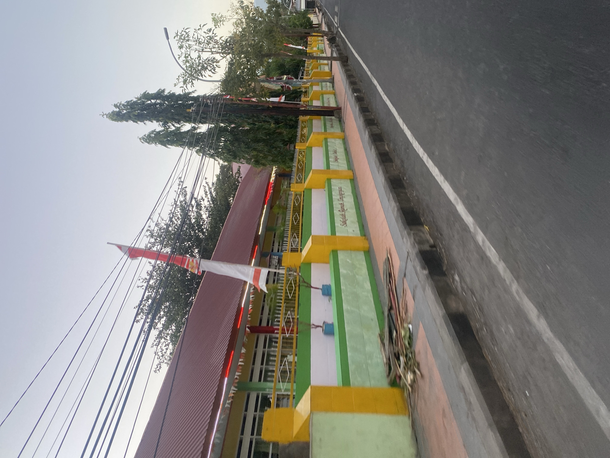 The image depicts a street scene in Bantaeng, Indonesia, with a school building visible on the left. The school has a red corrugated metal roof and yellow and green painted walls. A white and red Indonesian flag is flying on a pole in front of the school, and decorative pennant lights are strung along the roofline. A low, brightly colored wall with yellow posts separates the school property from the sidewalk and street. Trees line the sidewalk, and a street lamp is visible in the distance. The street itself is paved and has a white line marking its edge. Debris, including branches and leaves, is piled on the sidewalk near the wall. The sky is overcast, suggesting it is either early morning or late afternoon.