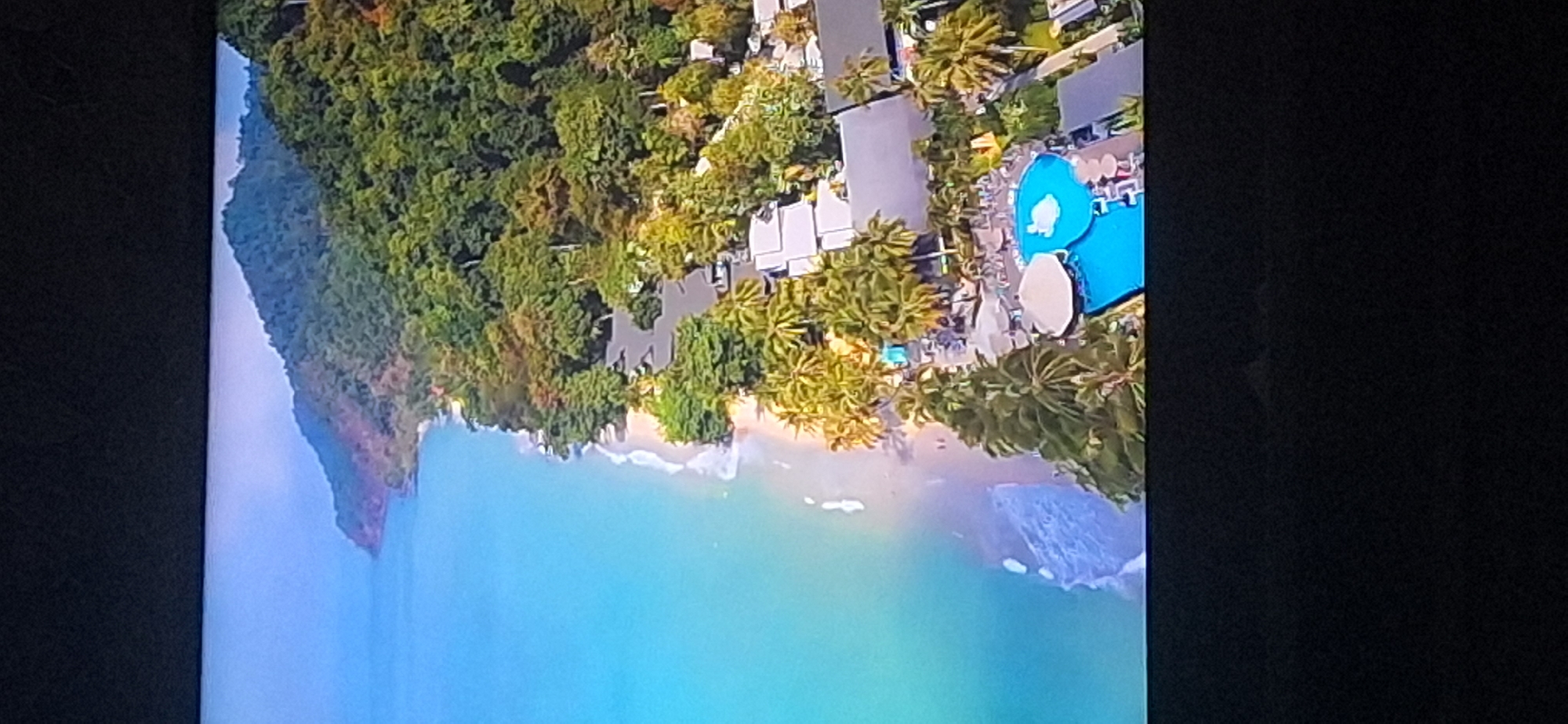 This is an aerial view of a tropical resort located on a coastline. The ocean is a vibrant turquoise and meets a sandy beach lined with palm trees. Behind the beach, a dense green forest covers rolling hills that lead up to a mountain in the distance. The resort itself features several buildings with white roofs and a prominent swimming pool with blue water and lounge chairs. There are a few people visible on the beach and near the pool area. This image does not depict any location in Bila Tserkva, Ukraine, as it is a tropical coastal scene.