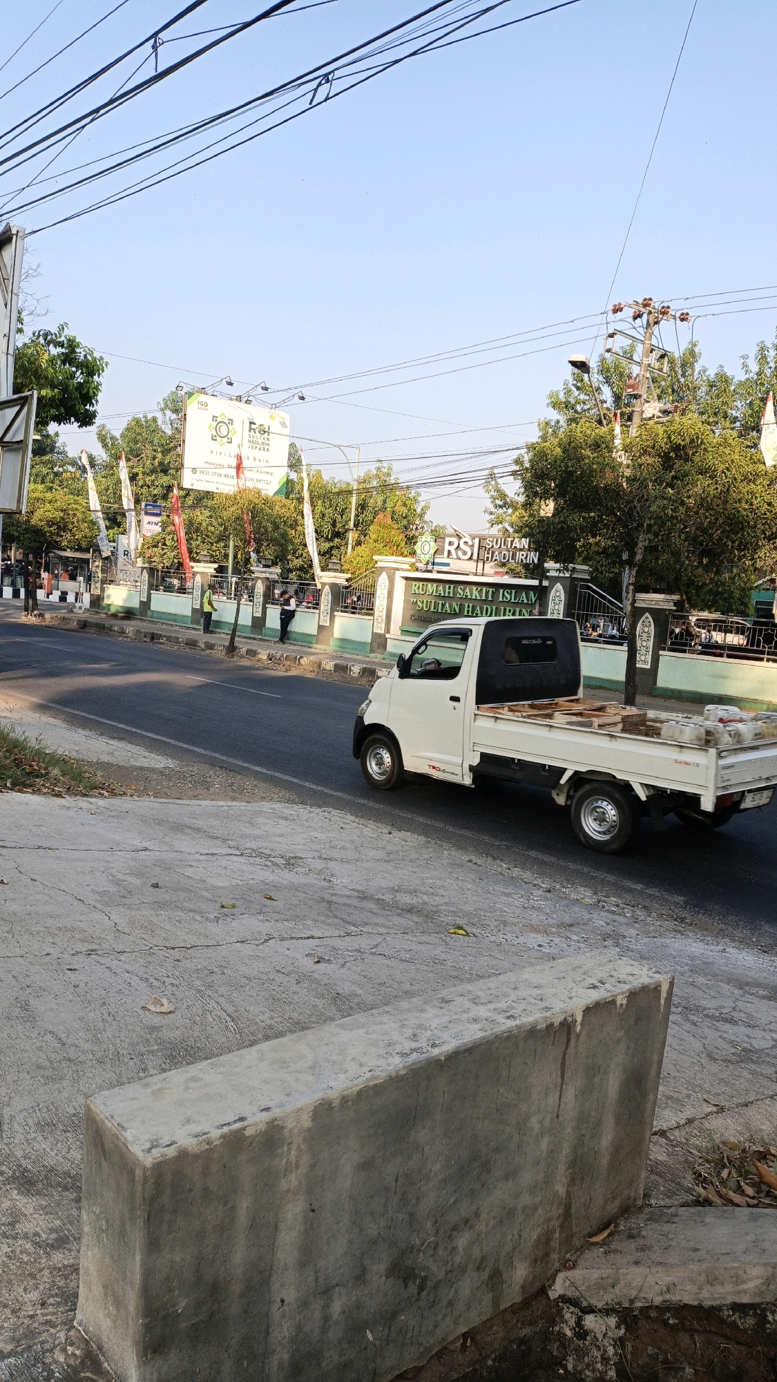 In the city of Kuwasen, Indonesia, a white pickup truck with a flatbed is seen driving on a paved road. The truck is loaded with what appear to be concrete blocks and wooden pallets. The scene takes place during the day, with a clear blue sky and sunlight casting shadows.
To the left of the road, there is a sidewalk with a concrete barrier in the foreground. Behind the sidewalk, there is a building with a sign that reads "RUMAH SAKIT ISLAM 'SULTAN HADLIRIN' JEPARA". Several people are visible standing near the building's entrance, and some are walking along the sidewalk. There are also trees and electrical poles lining the street.
The road itself is asphalt, with white lines marking the lanes. The environment suggests an urban or semi-urban setting, likely in a public area due to the presence of the hospital and street infrastructure. The overall impression is of a typical day in a bustling Indonesian city.