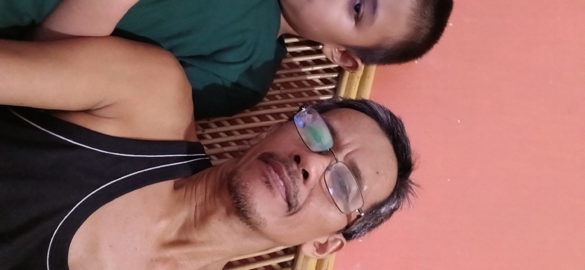 This is a close-up, selfie-style photograph of two individuals in what appears to be an indoor setting. On the right is an adult male with short, dark hair, graying at the temples, wearing clear-rimmed glasses. He has a thin mustache and goatee. He is wearing a black tank top with white trim. His left arm is visible. On the left, partially in the frame, is a younger individual, likely a child or adolescent, with short dark hair. This person is wearing a dark green t-shirt and their left ear is visible. Both individuals are positioned close to the camera, and the adult male's face is the primary focus. The background consists of a woven material, likely a headboard or partition, and a plain peach-colored wall. The lighting is somewhat dim, with reflections visible on the adult male's glasses. The photograph is likely taken in the city of Samarinda, Indonesia, given the context.