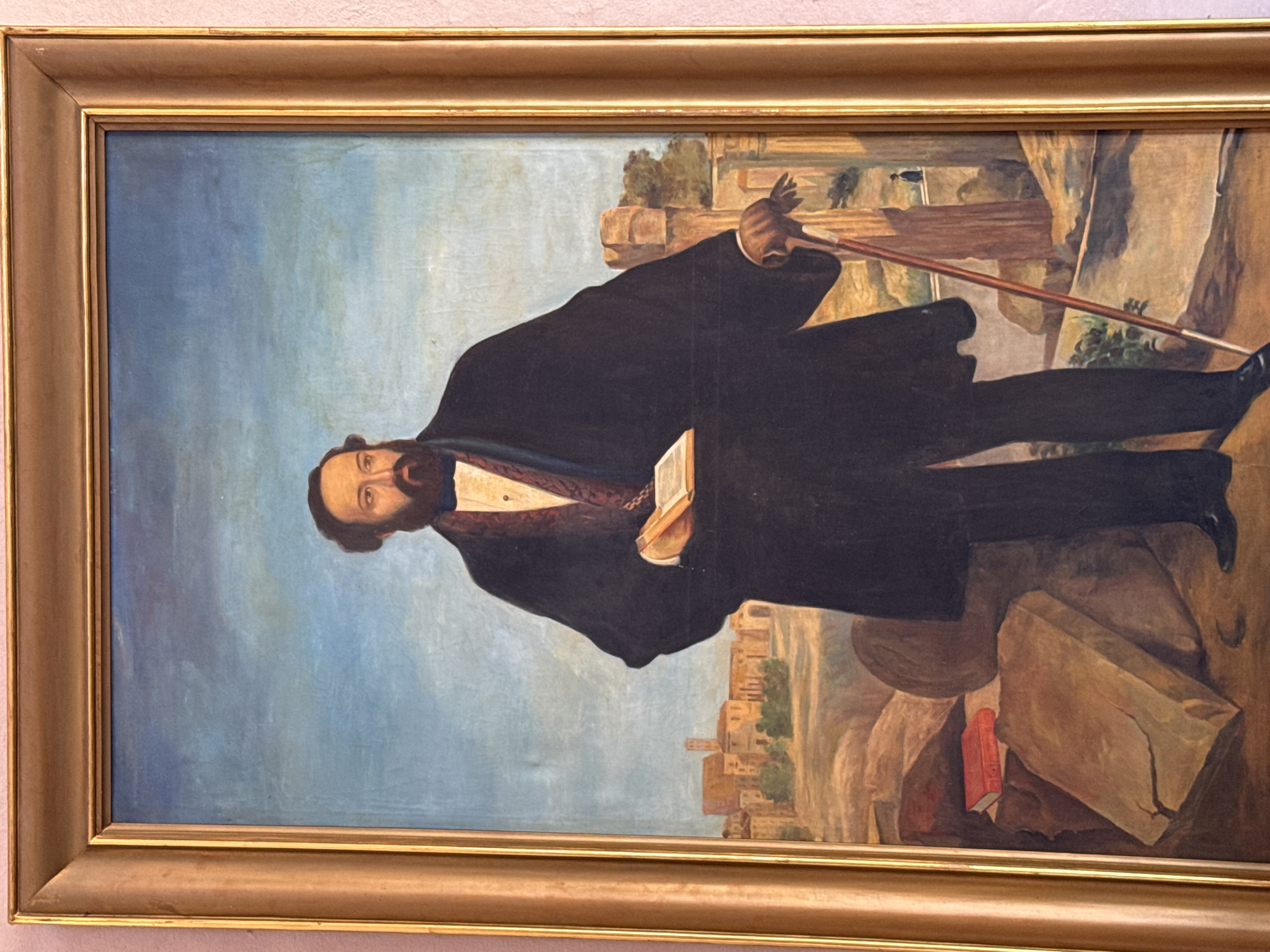 A framed portrait of a man with a beard and dark hair stands facing the viewer, angled slightly to the left. He wears a black coat, holding a book, and is accompanied by a walking stick. The painting is set against a landscape that includes architectural ruins and a distant town or city. It is unknown if it is in Querétaro, Mexico.