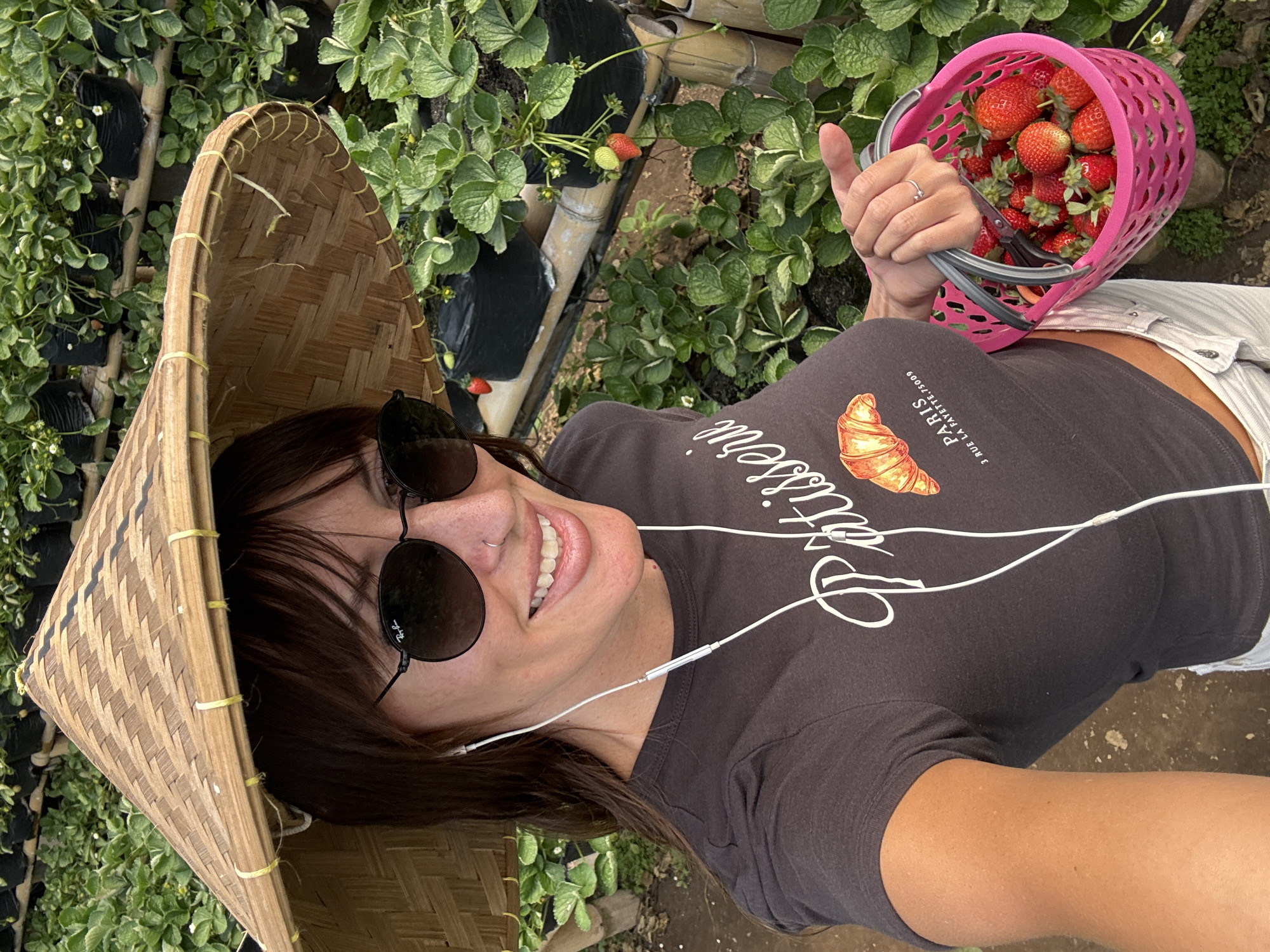 A medium shot shows a woman wearing a traditional conical hat and sunglasses, smiling at the camera. She is in a strawberry farm, holding a pink basket full of strawberries. The background is filled with rows of strawberry plants in black pots, supported by bamboo structures. The woman is wearing a dark grey t-shirt with a graphic of a croissant and the text "PATISSERIE PARIS 3 RUE LA FAYETTE, 75009". She is also wearing white headphones. The scene is outdoors, with natural lighting. The location is identified as a strawberry farm in Sukasada, Bali, Indonesia.
