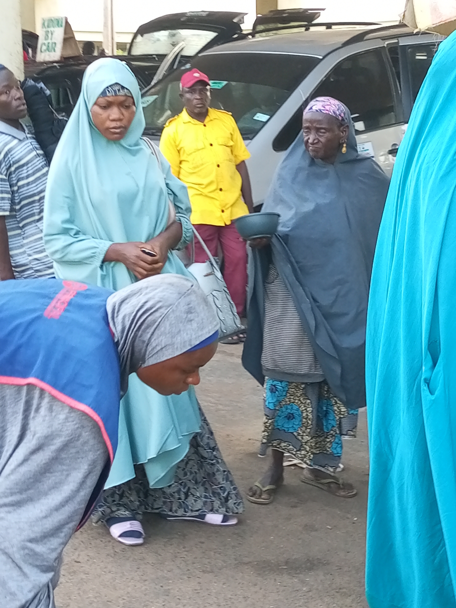 The image depicts a street scene in Gombe, Nigeria. In the foreground, a person wearing a blue and pink vest and a grey headscarf is bent over, appearing to be engaged in an activity. To their right, a woman in a light blue hijab stands with her hands clasped. Behind her, an elderly woman wearing a dark grey headscarf and a patterned skirt holds a dark bowl in her left hand. She is also wearing gold earrings. Another individual in a bright yellow shirt and a red cap stands to the far right, partially obscured. In the background, several vehicles are visible, including what appears to be a silver SUV and a darker car, suggesting a public or busy area. A sign with the text "KUDUNA BY CAR" is partially visible on the left side of the image. The ground is paved with asphalt.