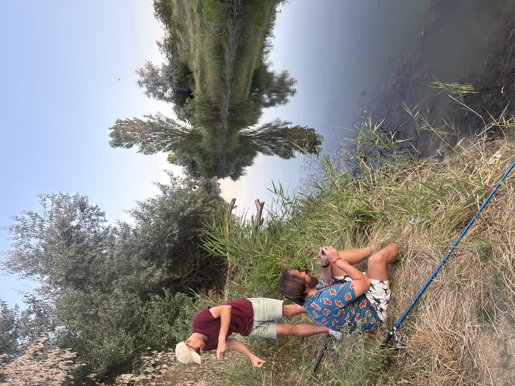 Two men are fishing on the bank of a calm river with dense vegetation on both sides. The man in the foreground, wearing a colorful short-sleeved shirt with a pattern of red and yellow shapes and patterned shorts, is sitting down and holding a fishing rod. The man in the background, standing and wearing a maroon t-shirt, light-colored shorts, and a beige cap, appears to be attending to his fishing line. The water reflects the trees and the clear sky above. The overall setting suggests a natural, tranquil environment on the outskirts of Krasnoflotskoye Rural Settlement, Ukraine.