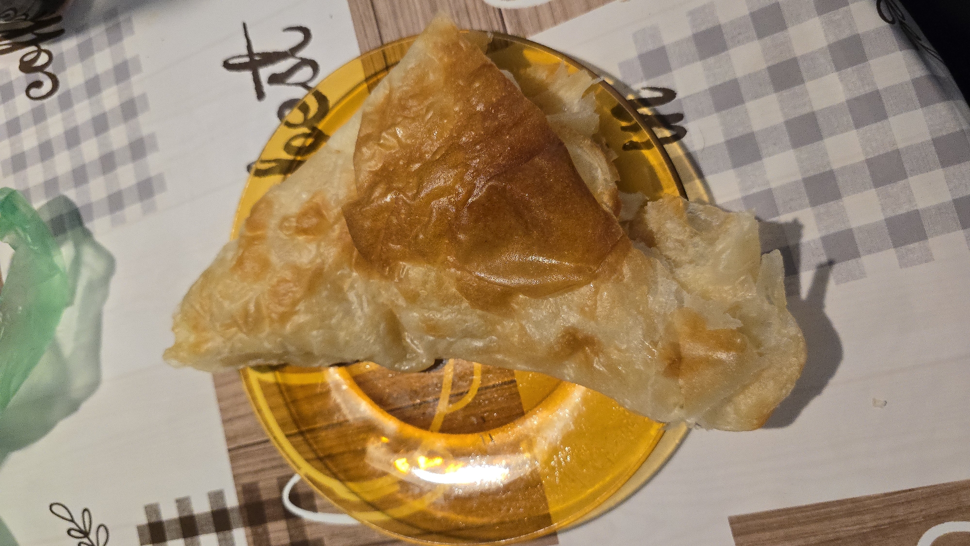 The image shows a close-up view of a pastry on a yellow plate. The pastry appears to be flaky and golden brown, with some layers visible. The plate has a patterned design. The pastry is placed on a checkered tablecloth. In the background, a crumpled green plastic bag is visible. Text elements, such as "coffee" and "best", are partially visible on the tablecloth. The lighting suggests an indoor setting, possibly a kitchen or dining area. No people are visible in the image. The scene is static and appears to be a still life of food.